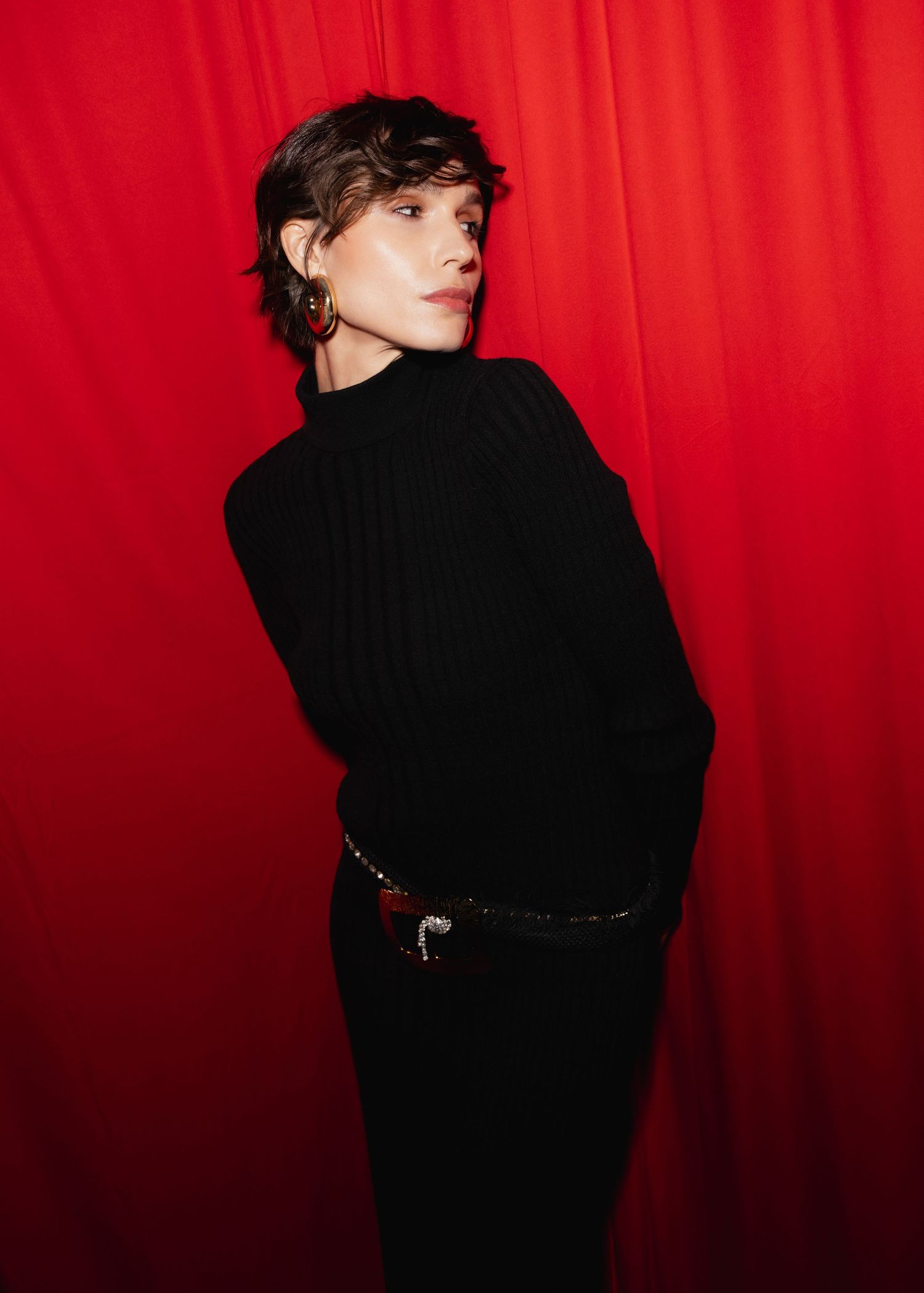 Artistic portrait against a vibrant red backdrop featuring a black turtleneck sweater and belt.