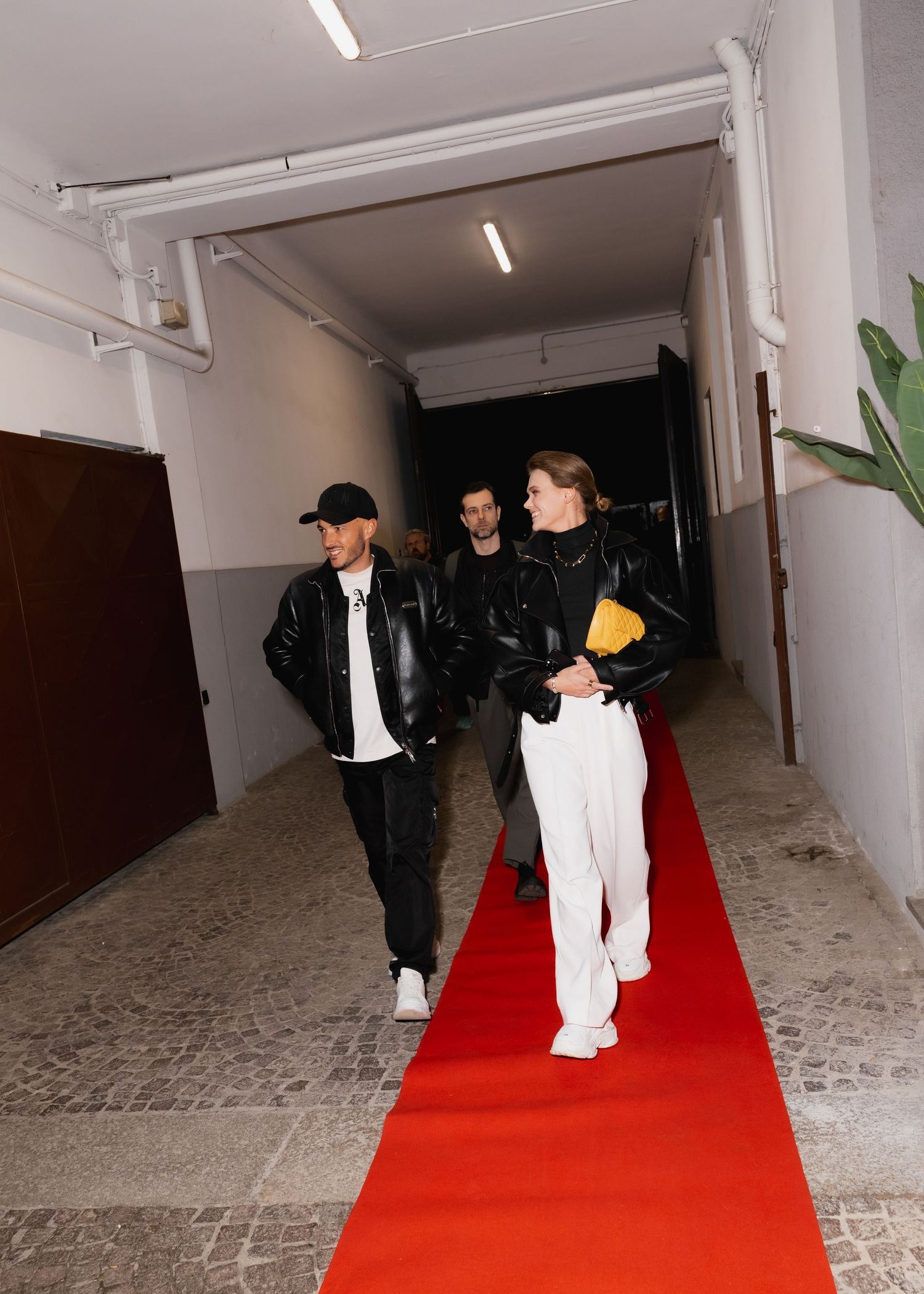 People walking on a red carpet in a white hallway with black doors and stone floor.