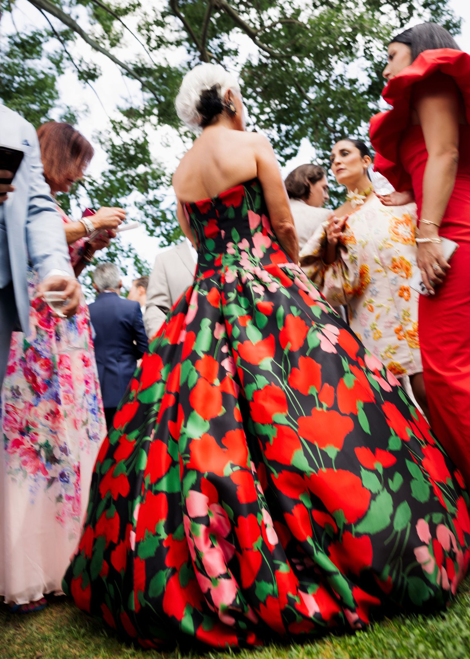Woman in a stunning red and green floral ball gown at an outdoor garden party event.