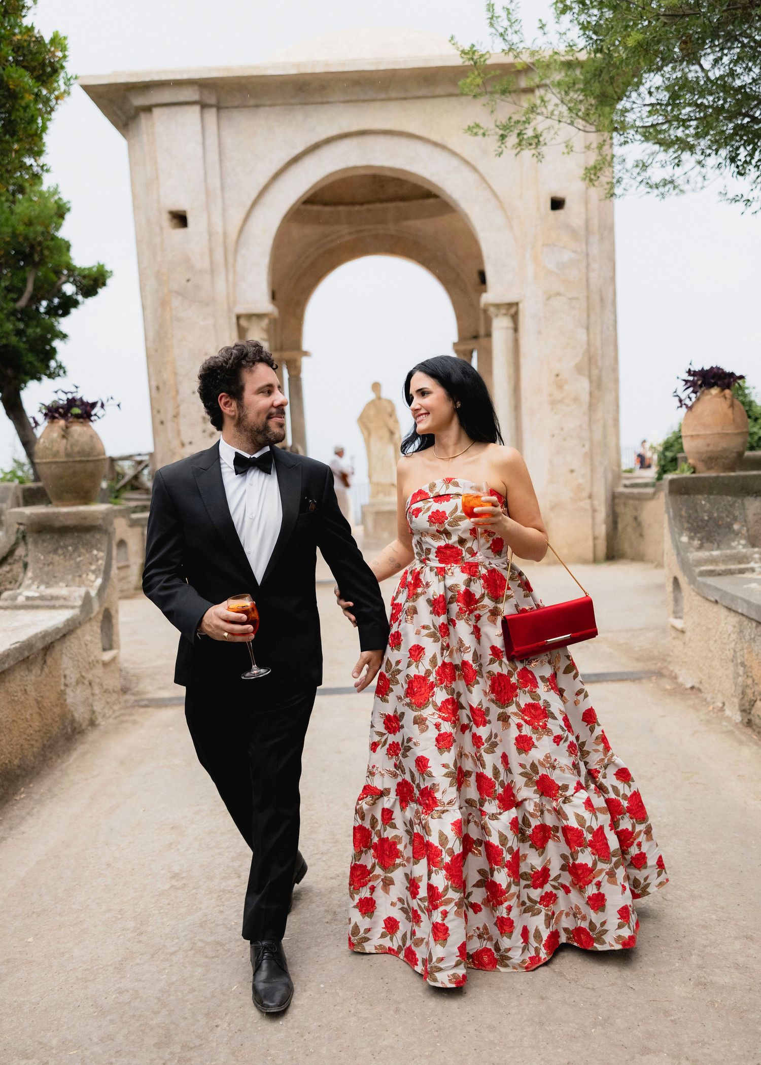 Couple in formal attire walking hand in hand near a stone arch, woman in floral gown, man in tuxedo, holding drinks.