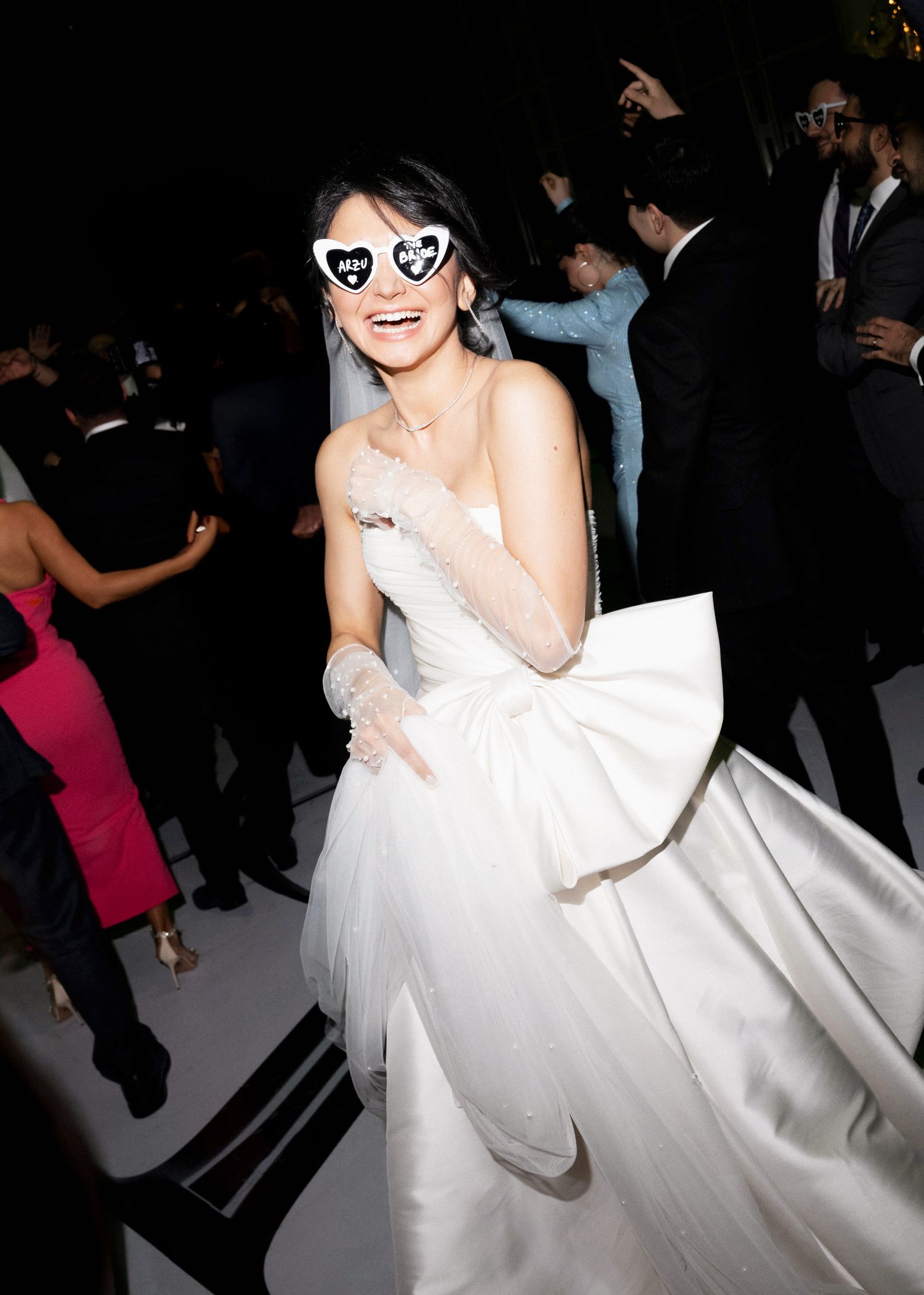 Woman in white strapless ballgown and heart-shaped sunglasses smiles at a formal event.