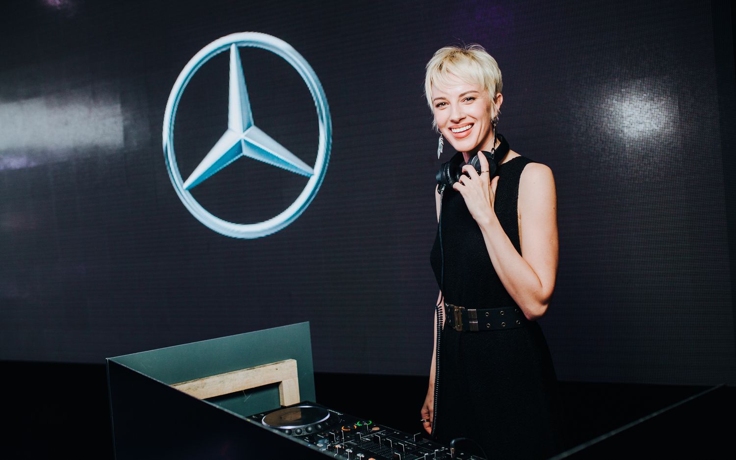 A DJ performs behind decks at an event with a large illuminated Mercedes-Benz logo on the black wall.