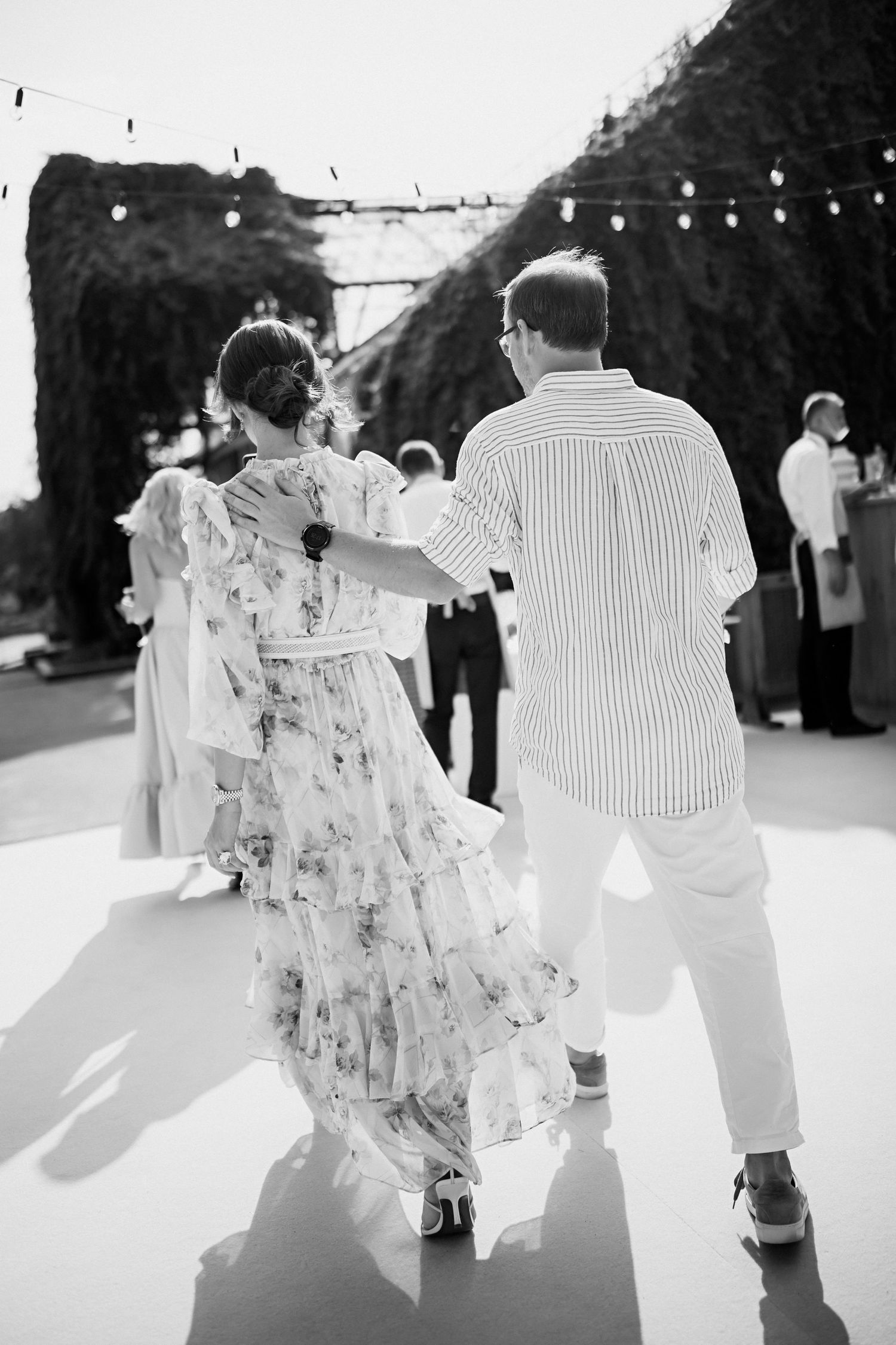 Black and white photo of couple dancing outdoors with string lights overhead.