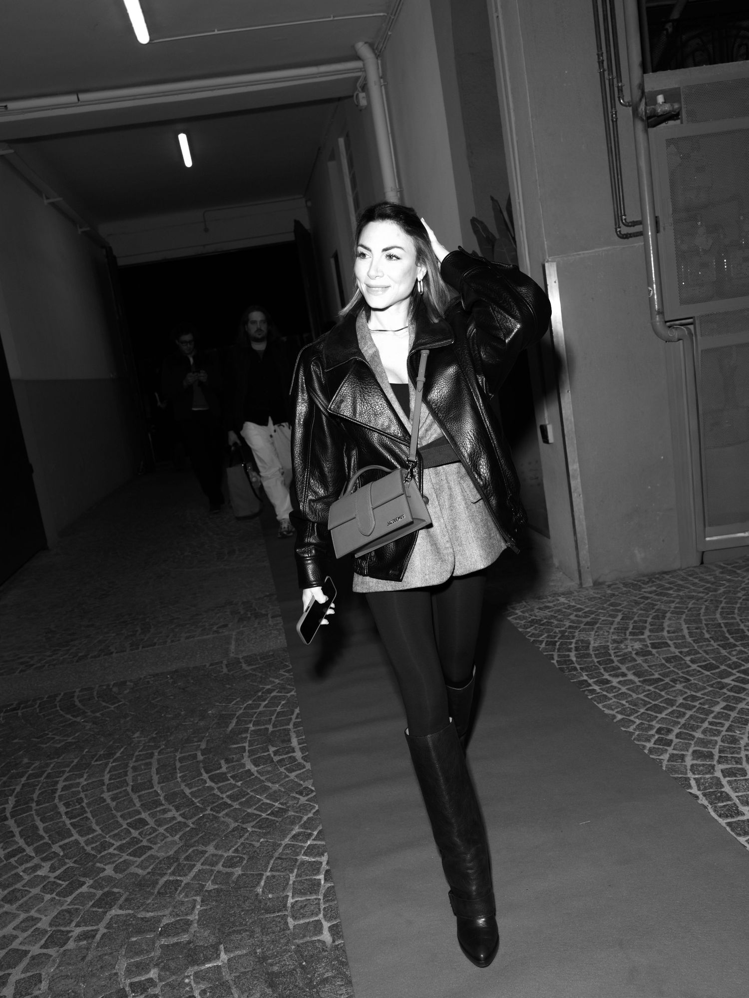 A person in a black leather jacket and dark pants walks through a dimly lit corridor with cobblestone flooring.