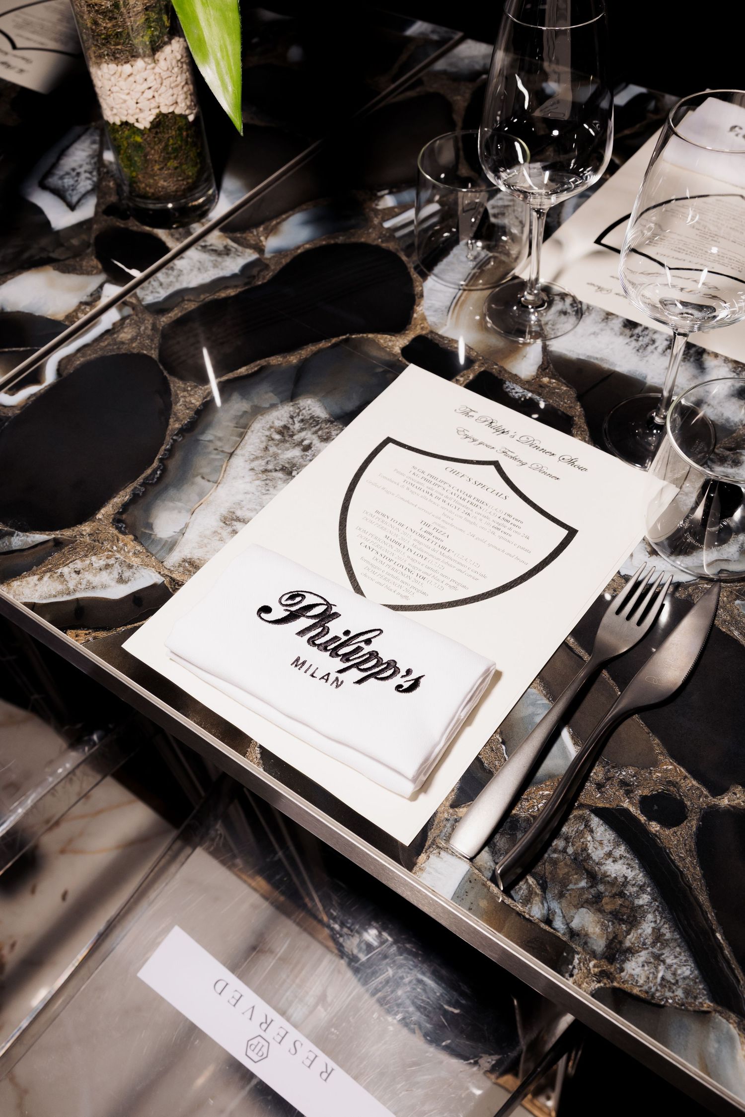 An elegant place setting features a menu card with a shield logo alongside wine glasses on a reflective table surface.