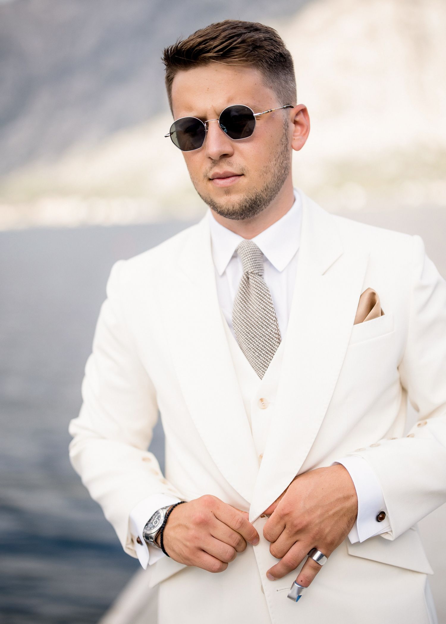 Stylish man in white suit and sunglasses adjusting jacket outdoors near water and mountains.