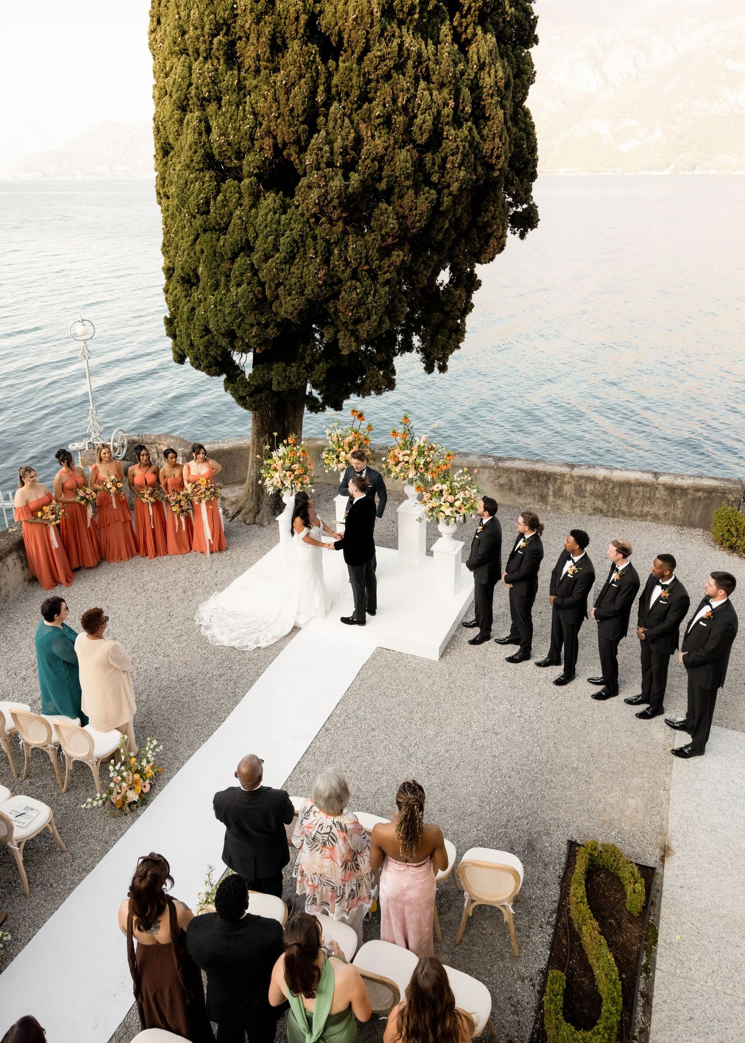 Aerial view of an elegant outdoor wedding ceremony on a lakeside terrace with bridesmaids in rust gowns and groomsmen in black suits.