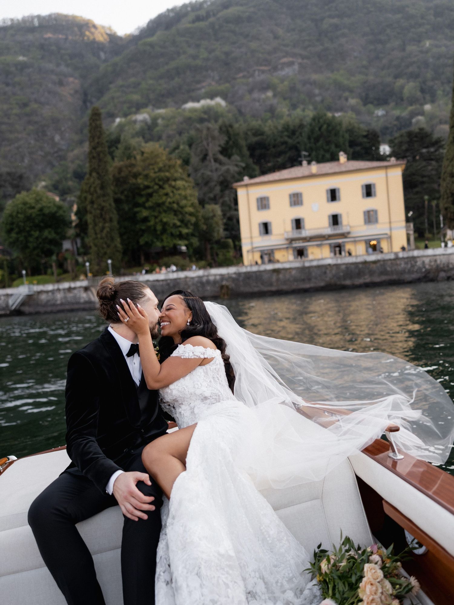 Romantic couple shares intimate moments in a wooden boat on a scenic lake with mountains and buildings in the background.