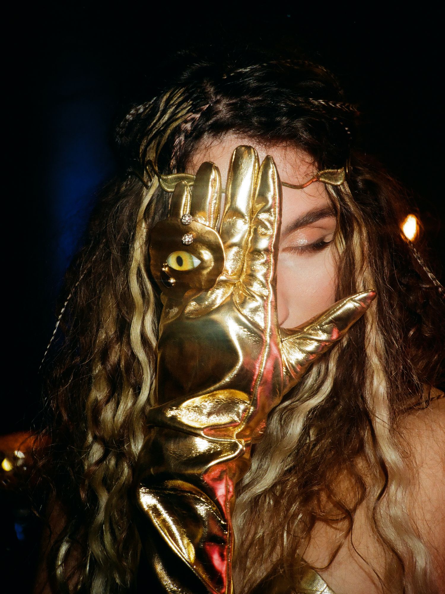 Close-up view of a performer wearing a metallic gold mask with long dark hair performing on stage in dramatic lighting.