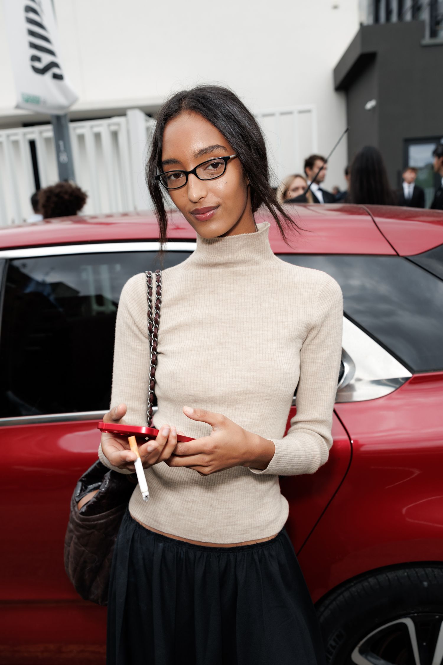 A person wearing a beige turtleneck sweater and black skirt stands next to a red car while using their phone.