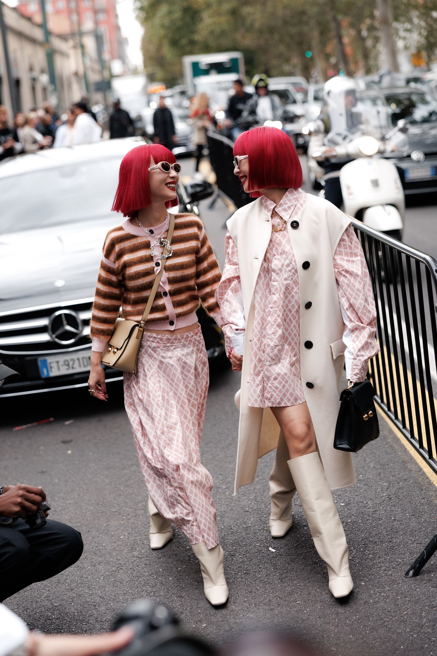 Two matching pink-haired fashionistas in stylish outfits with knee-high boots walk together on a city street.
