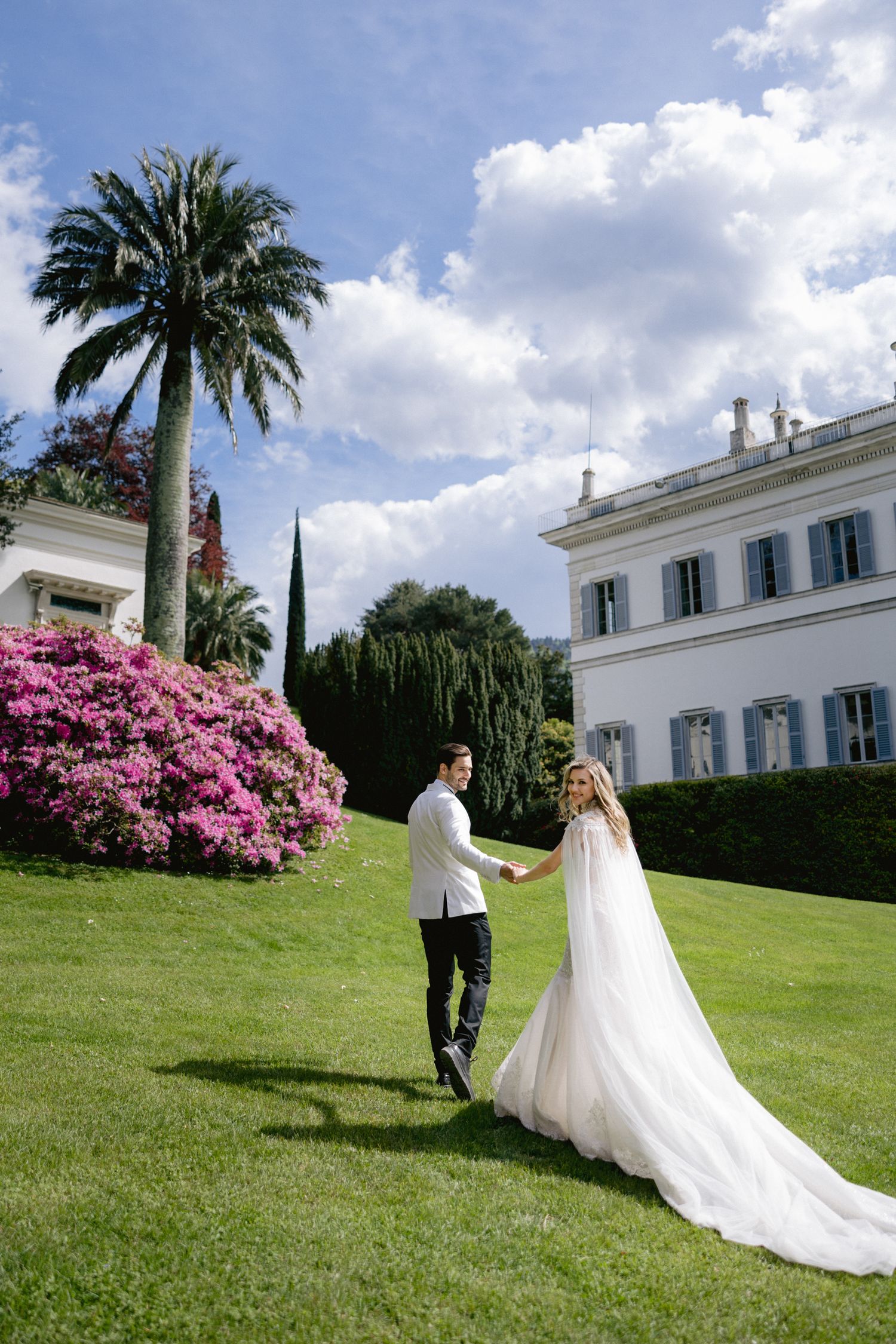 Newlyweds walk hand in hand on luxurious estate grounds with blooming azaleas and palm trees.