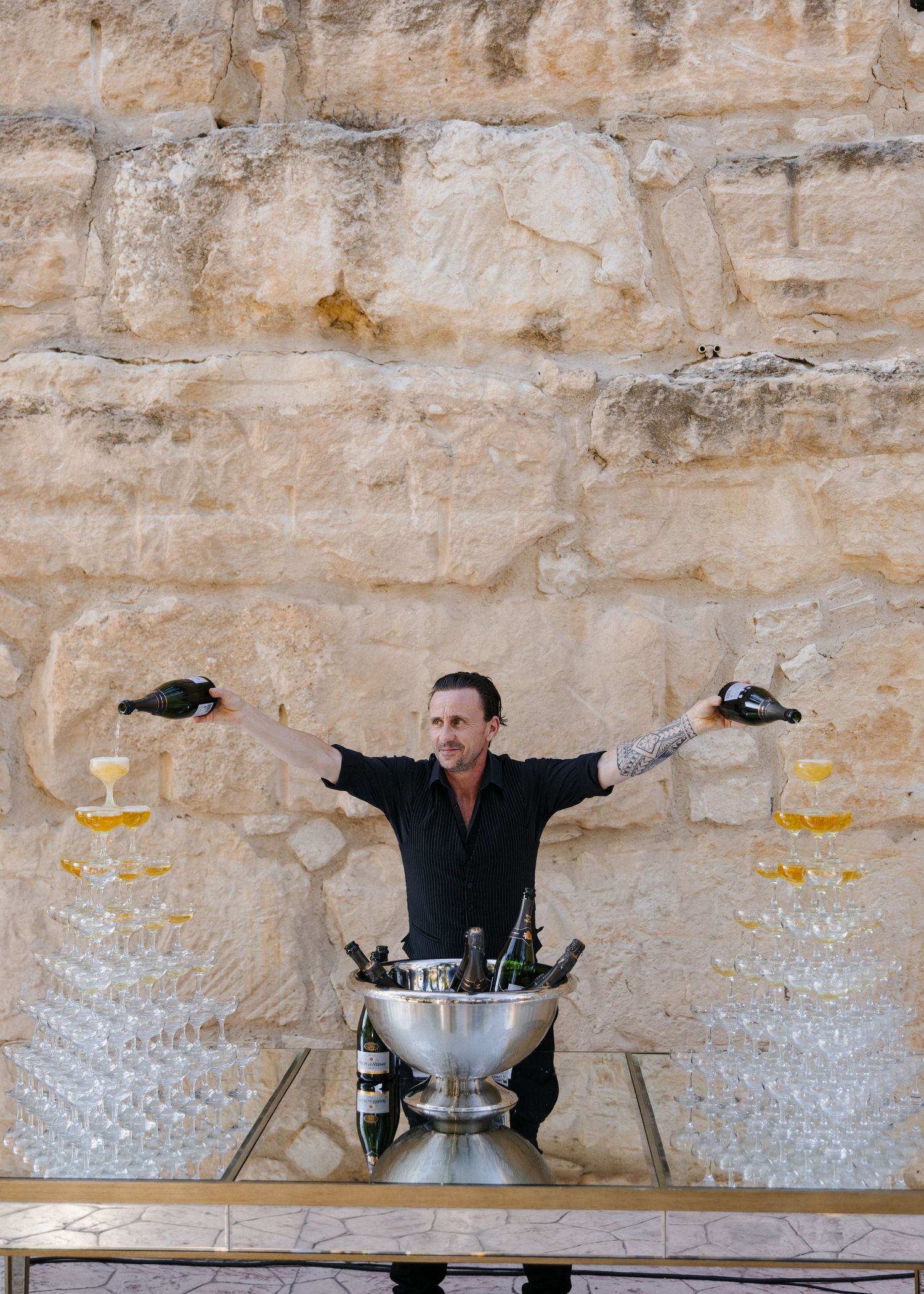 A bartender poses dramatically with champagne glasses against a limestone wall backdrop.