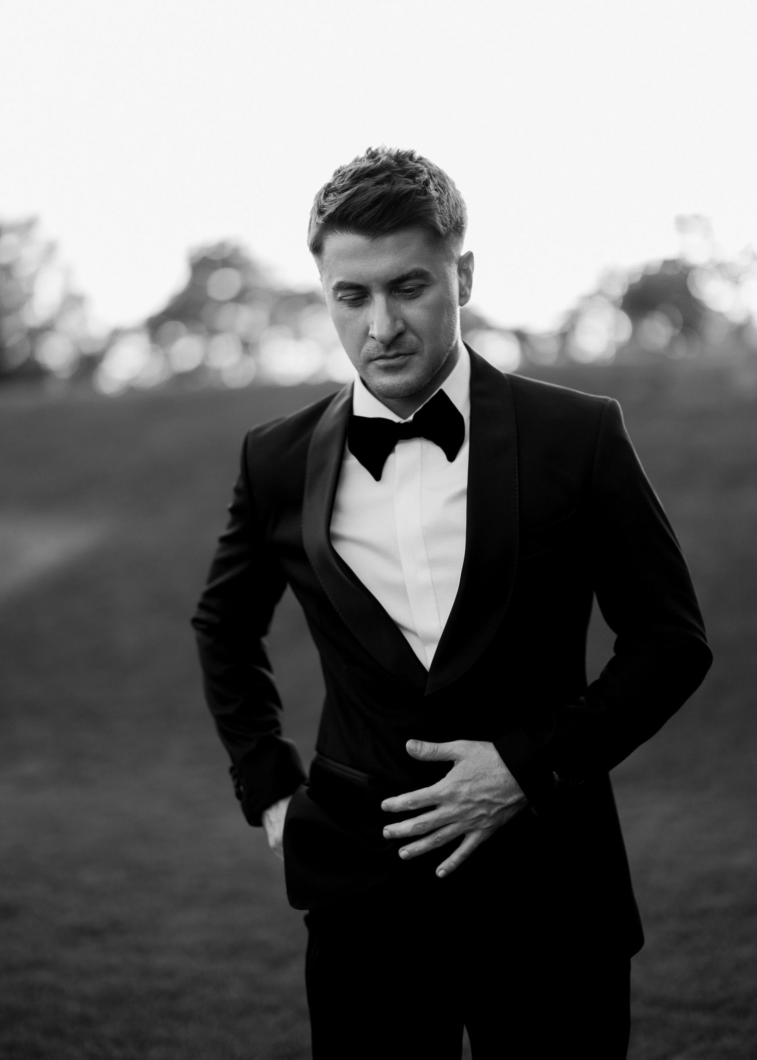 Man in black tuxedo with bow tie standing outdoors in black and white fashion portrait photo.