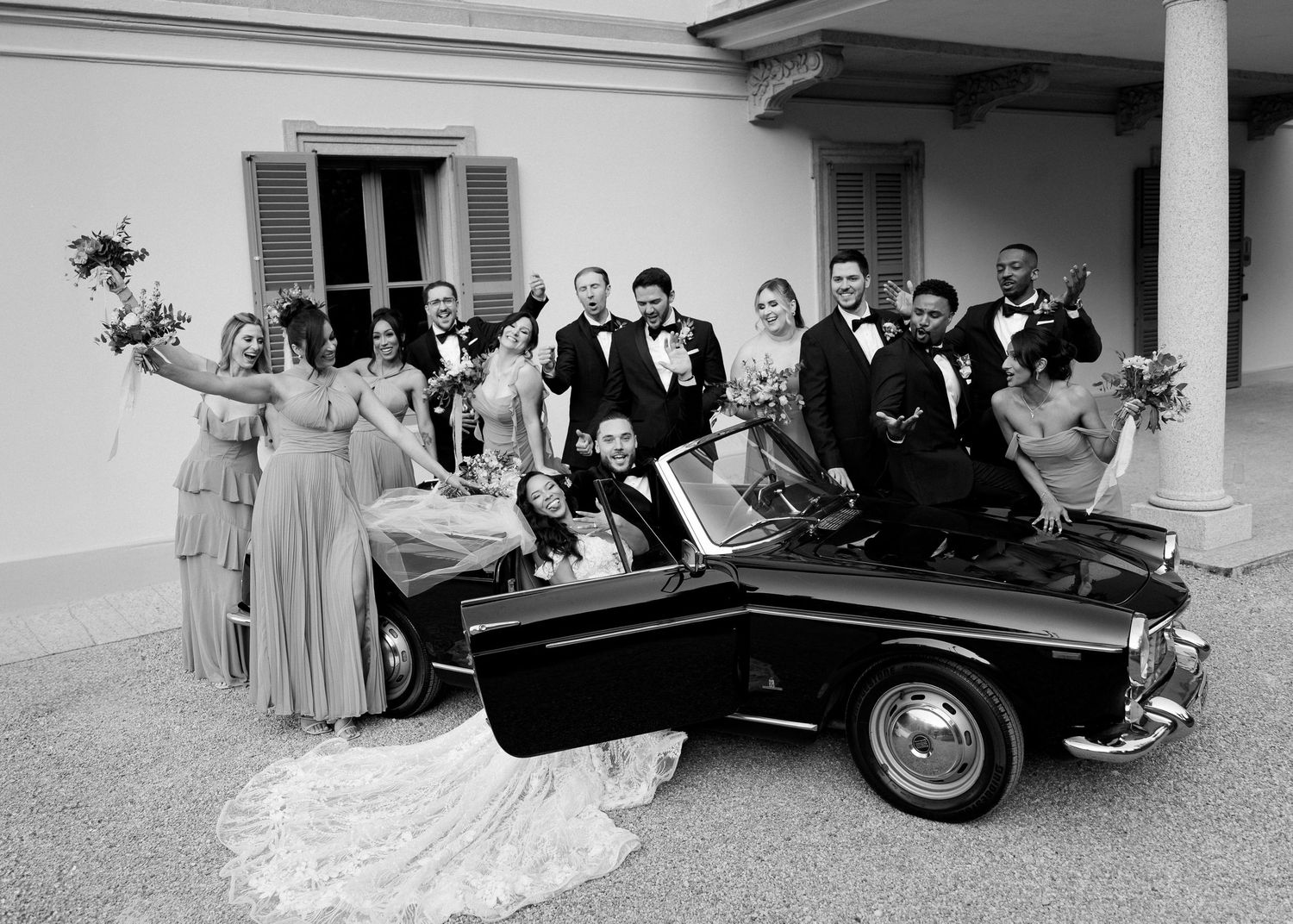 Black and white wedding party photo with bride, groom, and guests posing around a vintage convertible car.