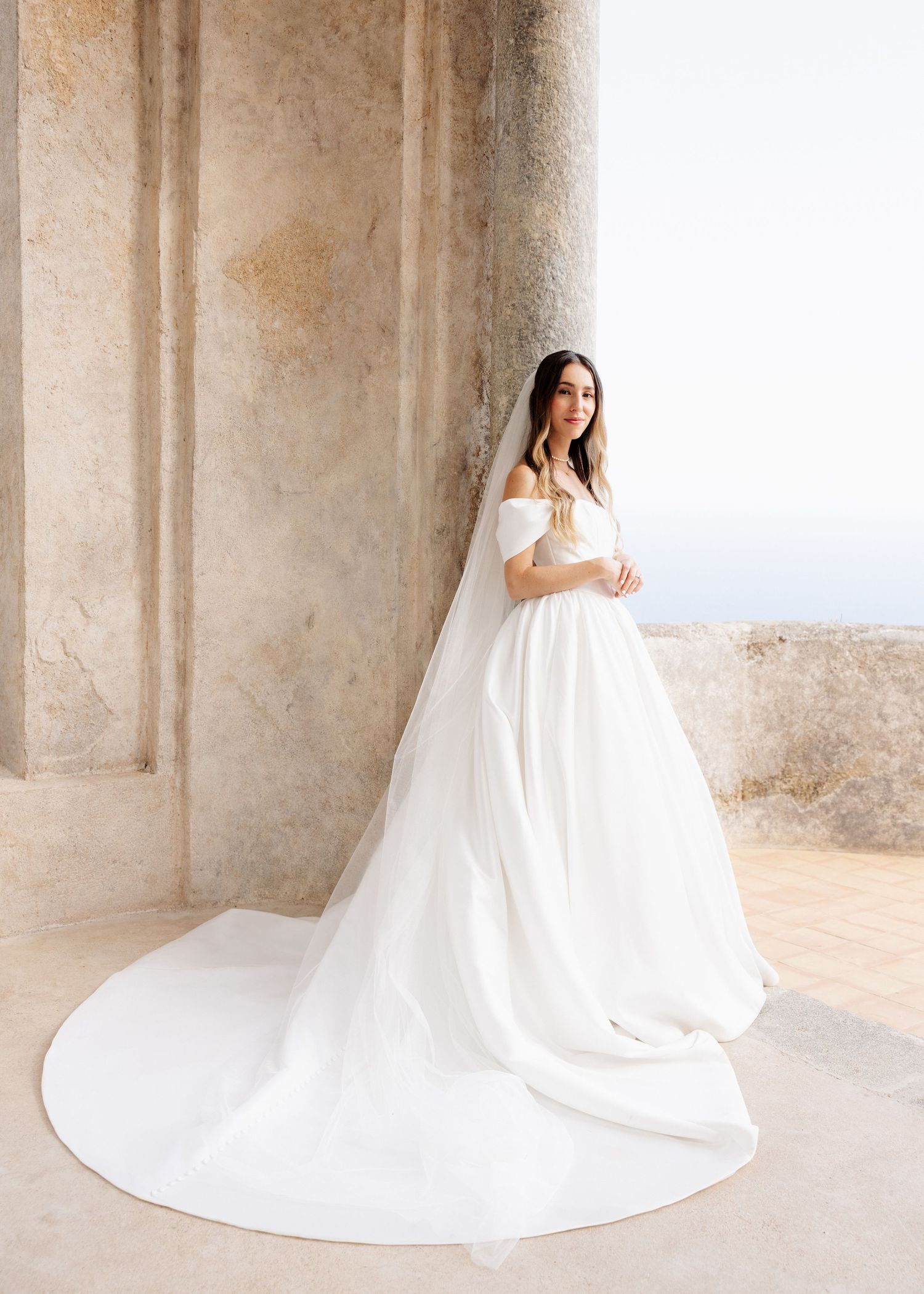 Bride in off-shoulder white ball gown with cathedral train and veil, posing by stone columns.
