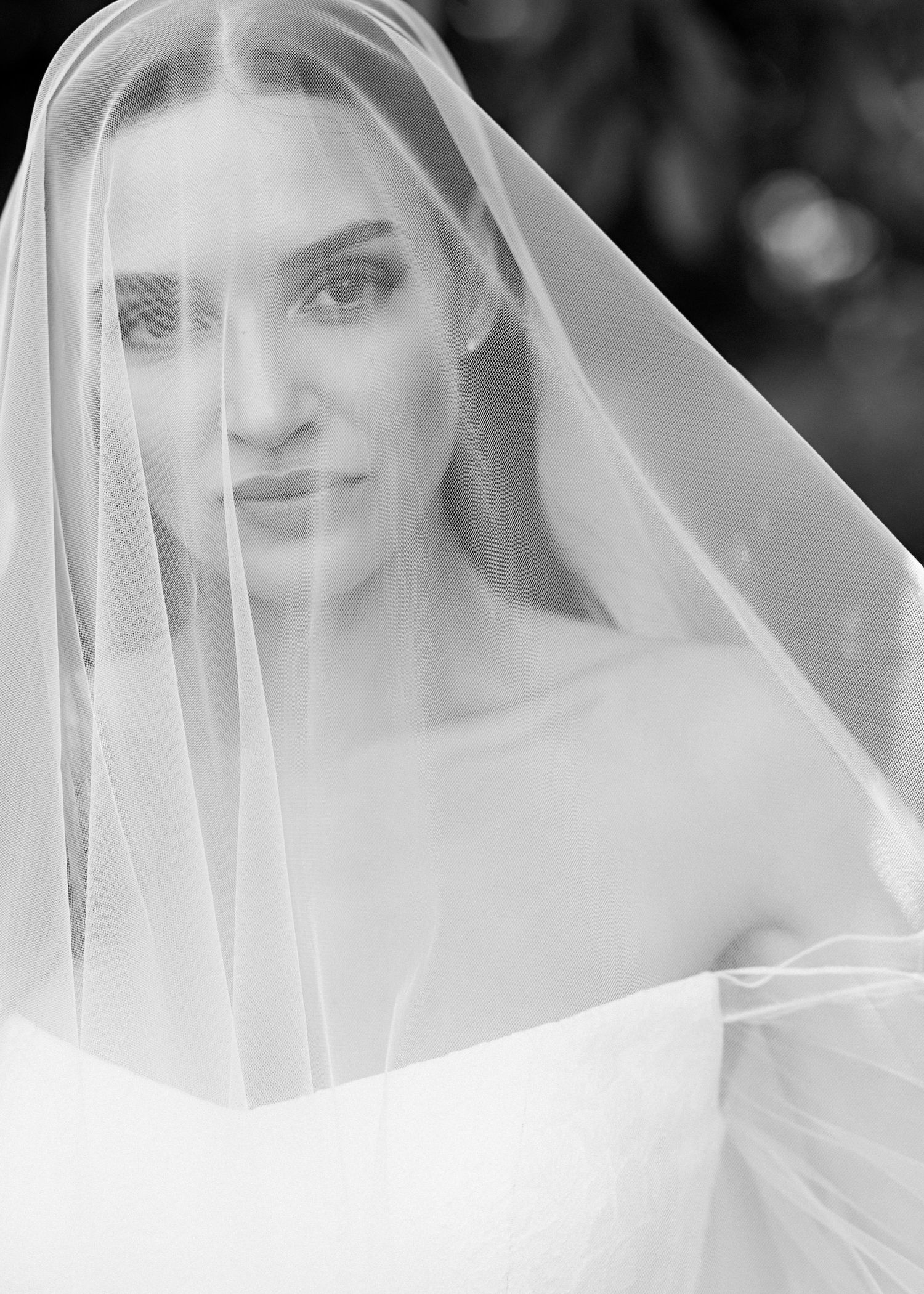 Black and white bridal portrait of a blonde woman wearing a flowing veil draped over her face in an elegant wedding gown.