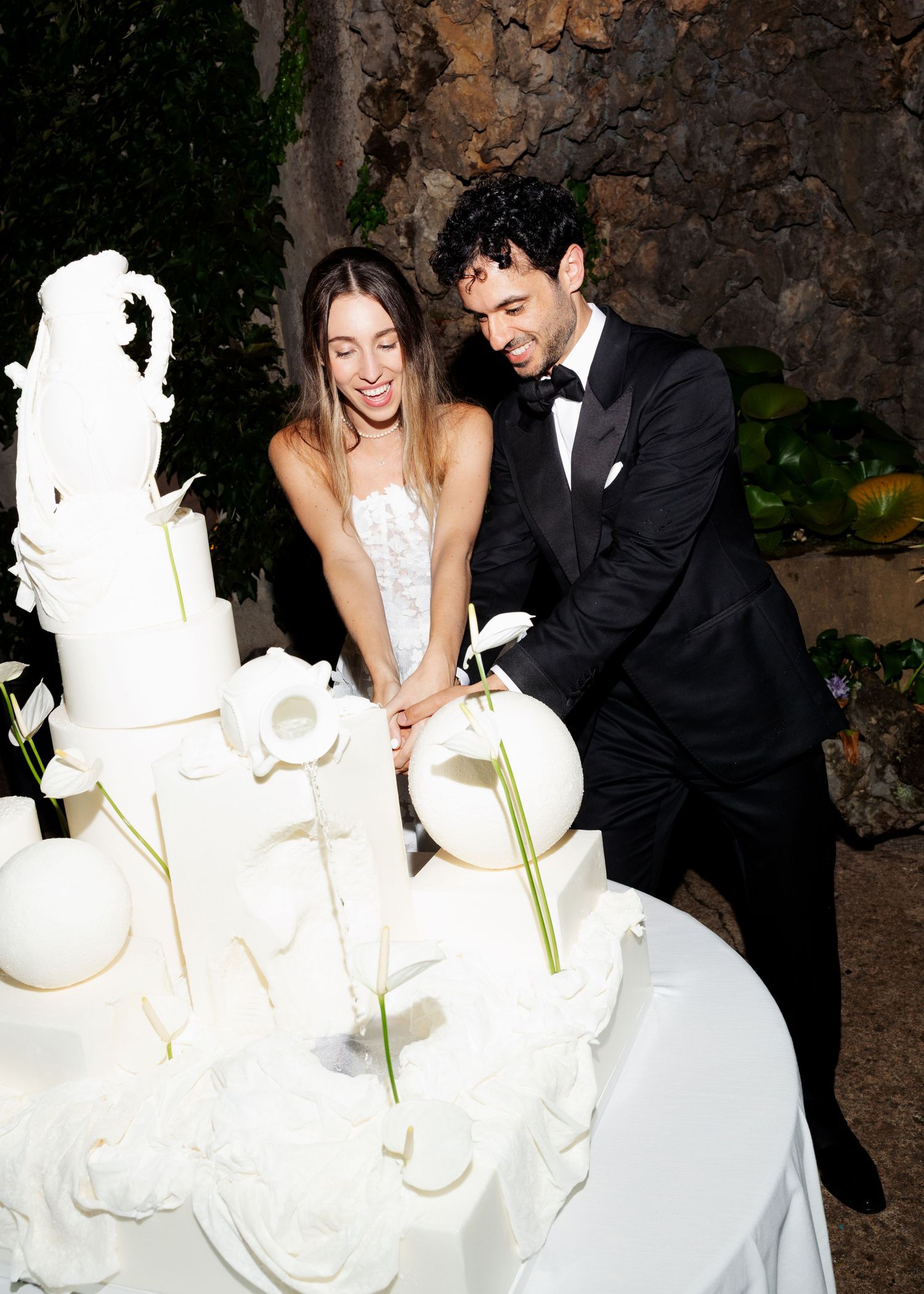 Bride and groom cutting a white sculptural wedding cake together, dressed in wedding attire at an elegant reception.