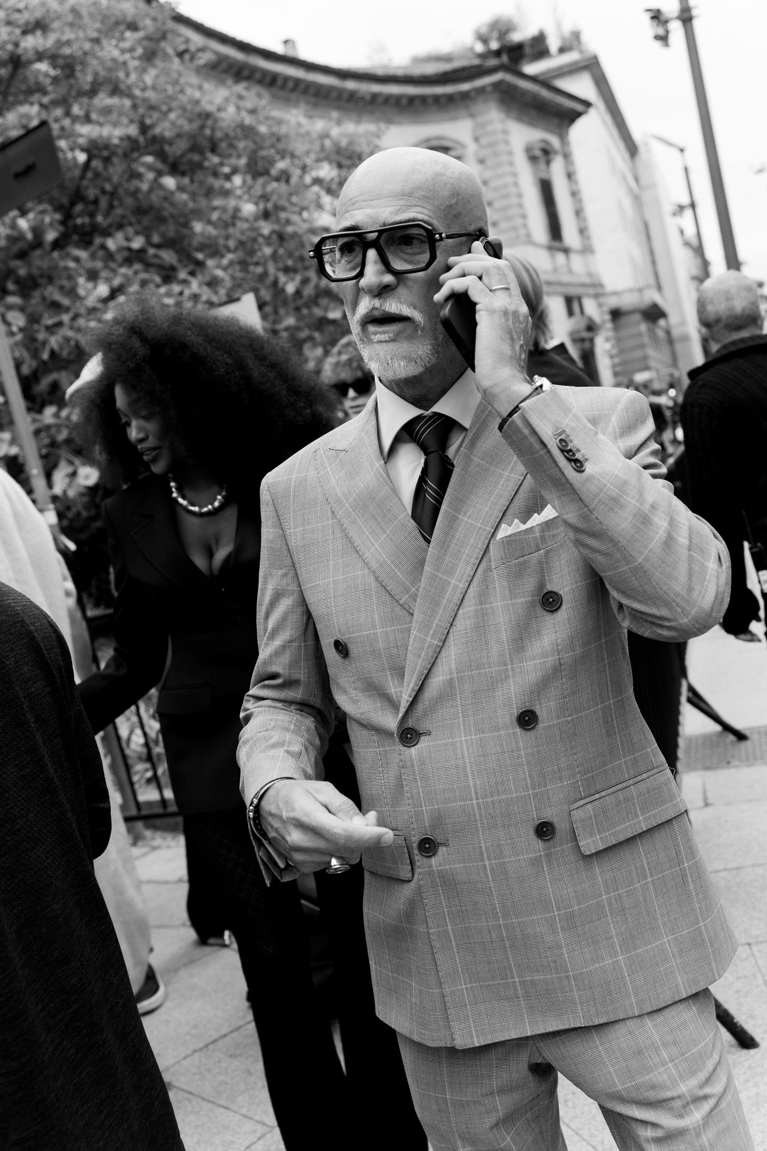 A well-dressed businessman in a plaid double-breasted suit and glasses speaks on a mobile phone outdoors.