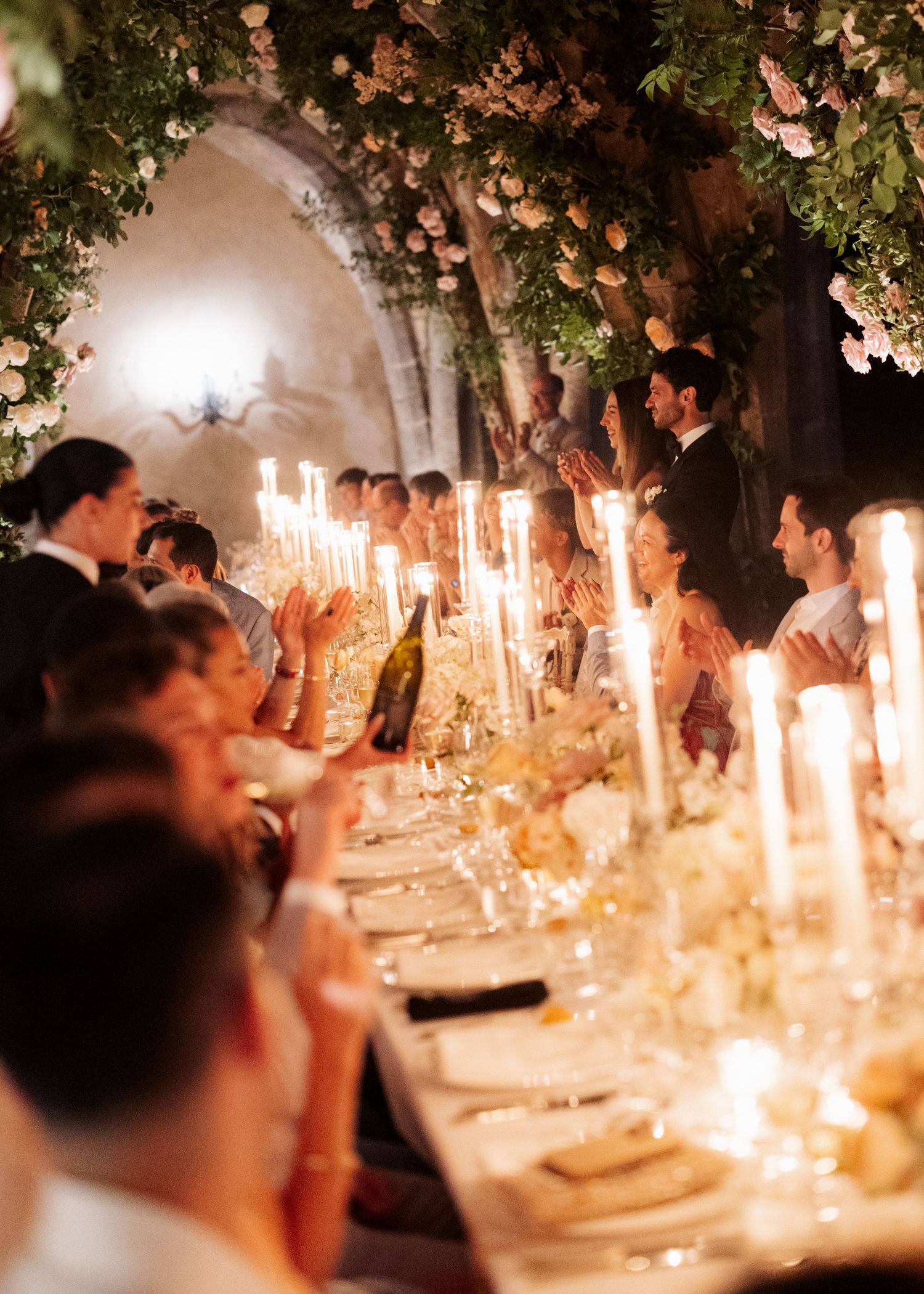 Elegant candlelit wedding reception with tall tapers, floral arrangements, and guests seated at a long decorated table.