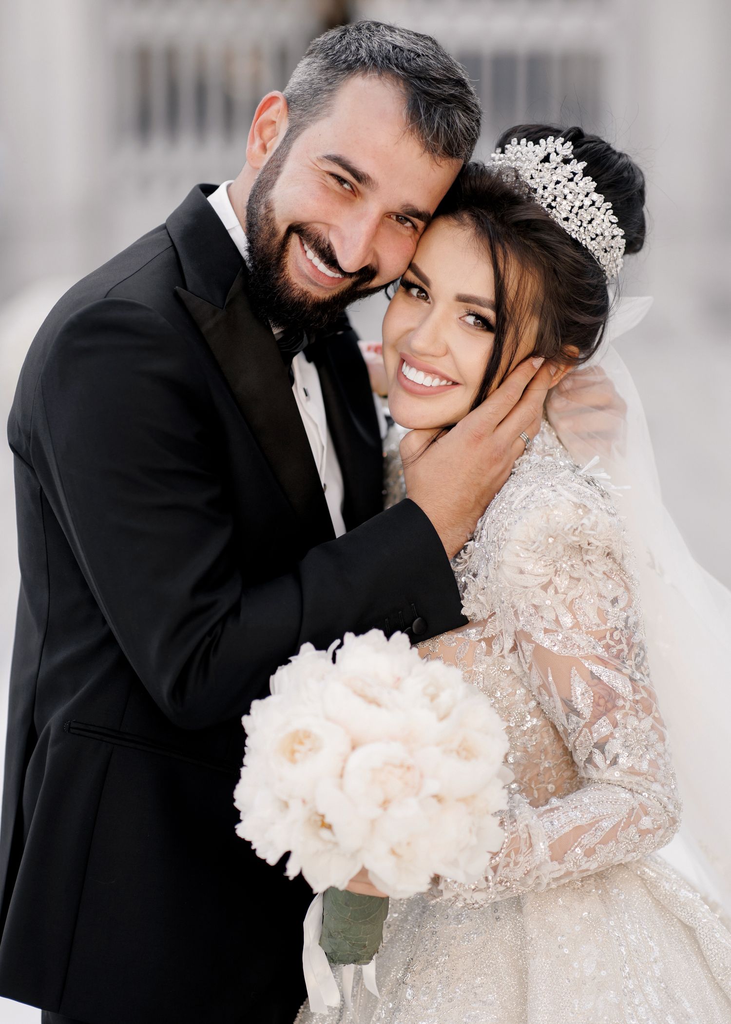 A smiling groom in a black tuxedo embraces his bride wearing a lace gown, tiara, and holding white peony bouquet.