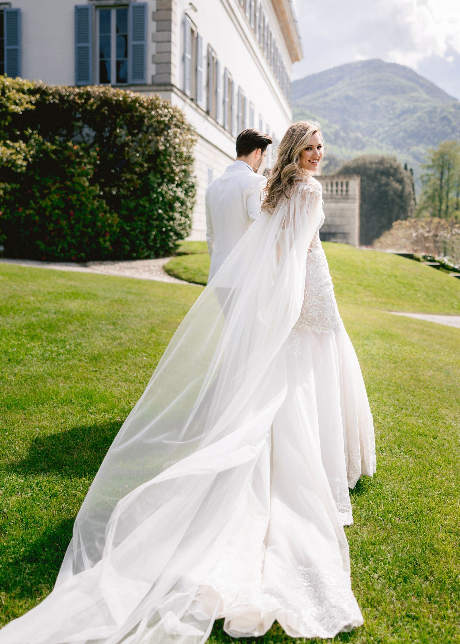 Bride in a flowing white gown and long veil smiles over her shoulder with groom outdoors at an elegant villa.