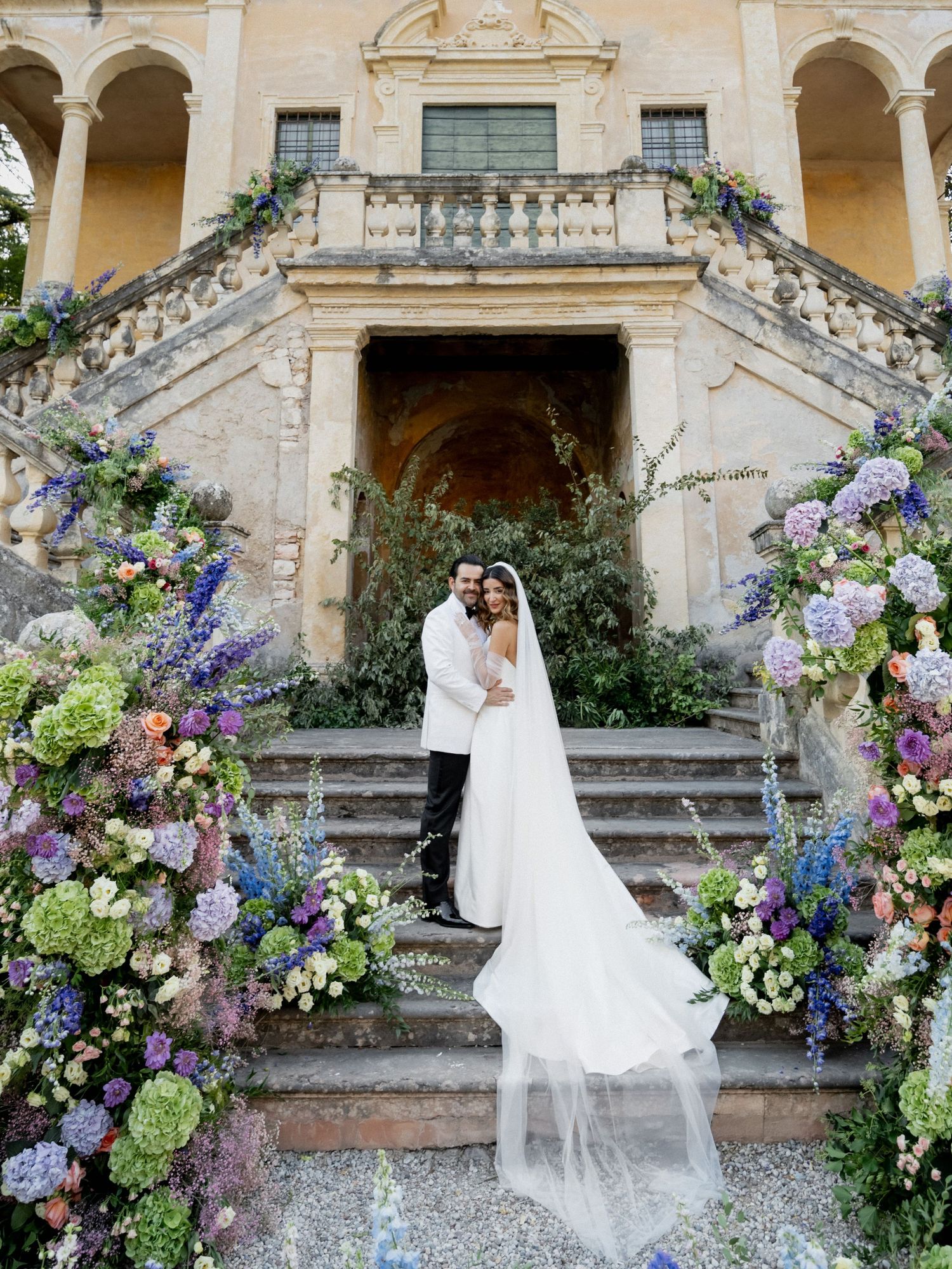 Wedding couple on stone steps with elaborate purple and blue floral arrangements.
