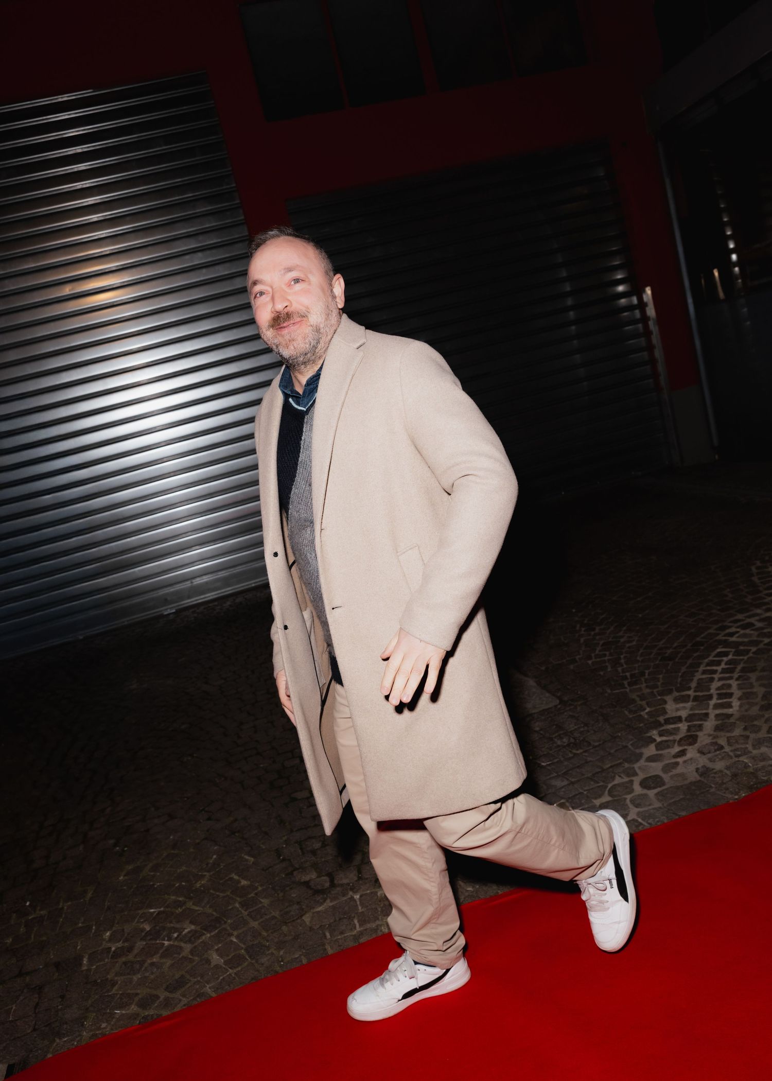 A person wearing a beige coat poses on a red carpet against a dark background with metal shutters.