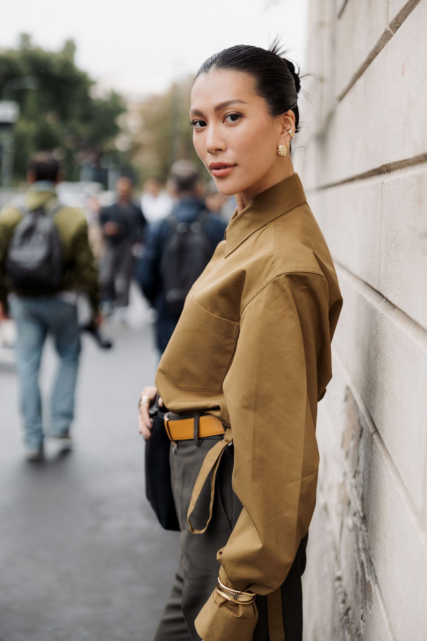 A person in a tan coat poses against a white wall in an urban setting with pedestrians in the background.