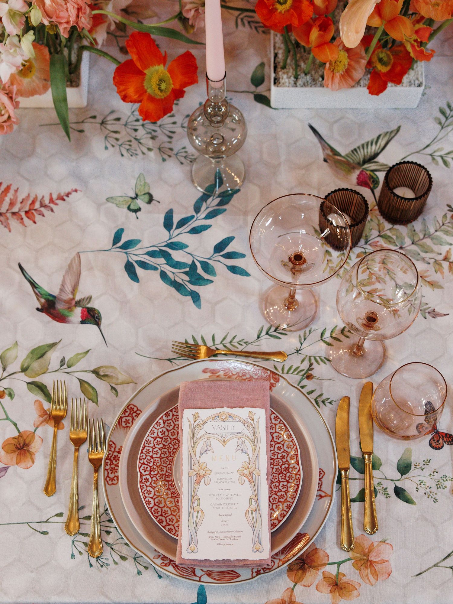 Wedding reception table setting with floral centerpieces, gold cutlery, and pink patterned plates on floral tablecloth.