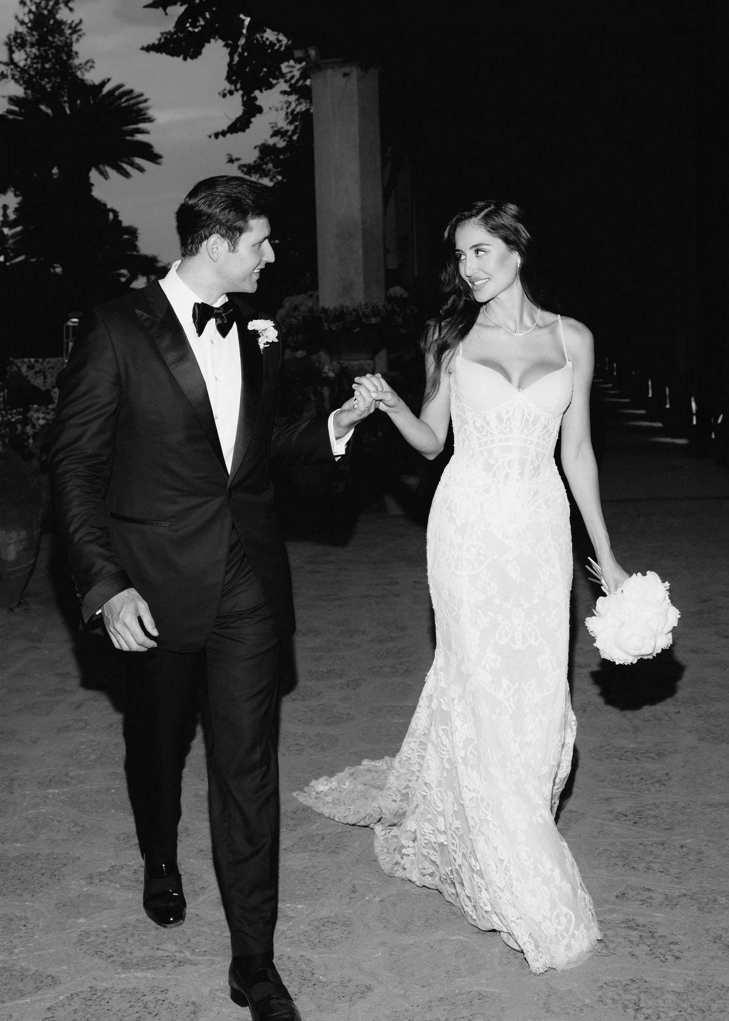 Black and white wedding photo of bride in lace gown holding bouquet and groom in tuxedo walking hand in hand.