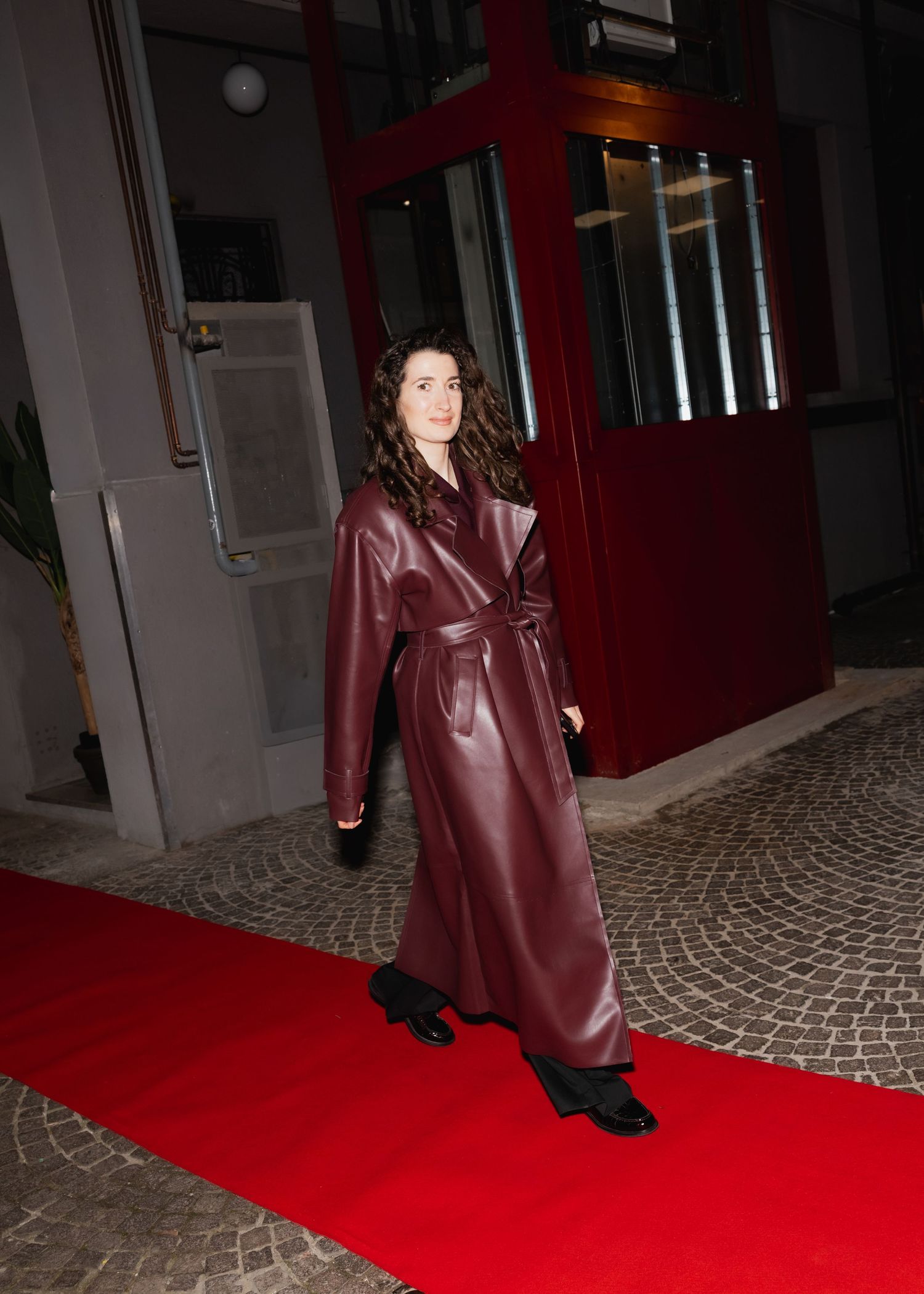 A model walks on a red carpet wearing a burgundy leather trench coat and black shoes in a dimly lit setting.