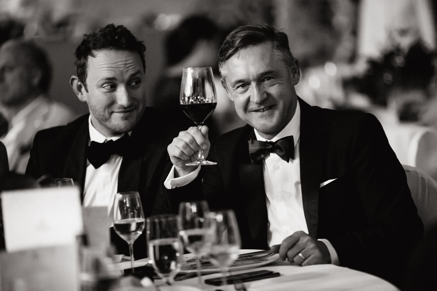 Classic black and white photo of two men in tuxedos toasting with wine glasses at formal dinner.