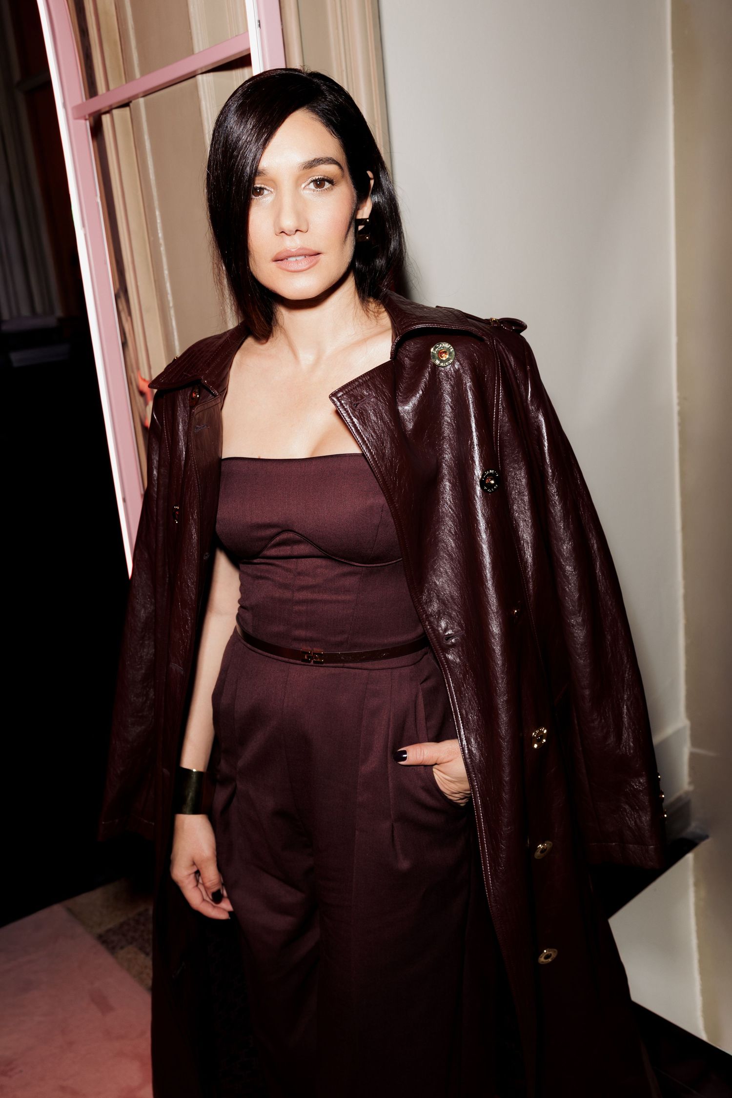 A person wearing a burgundy dress and dark leather jacket poses in a hallway against a white wall.