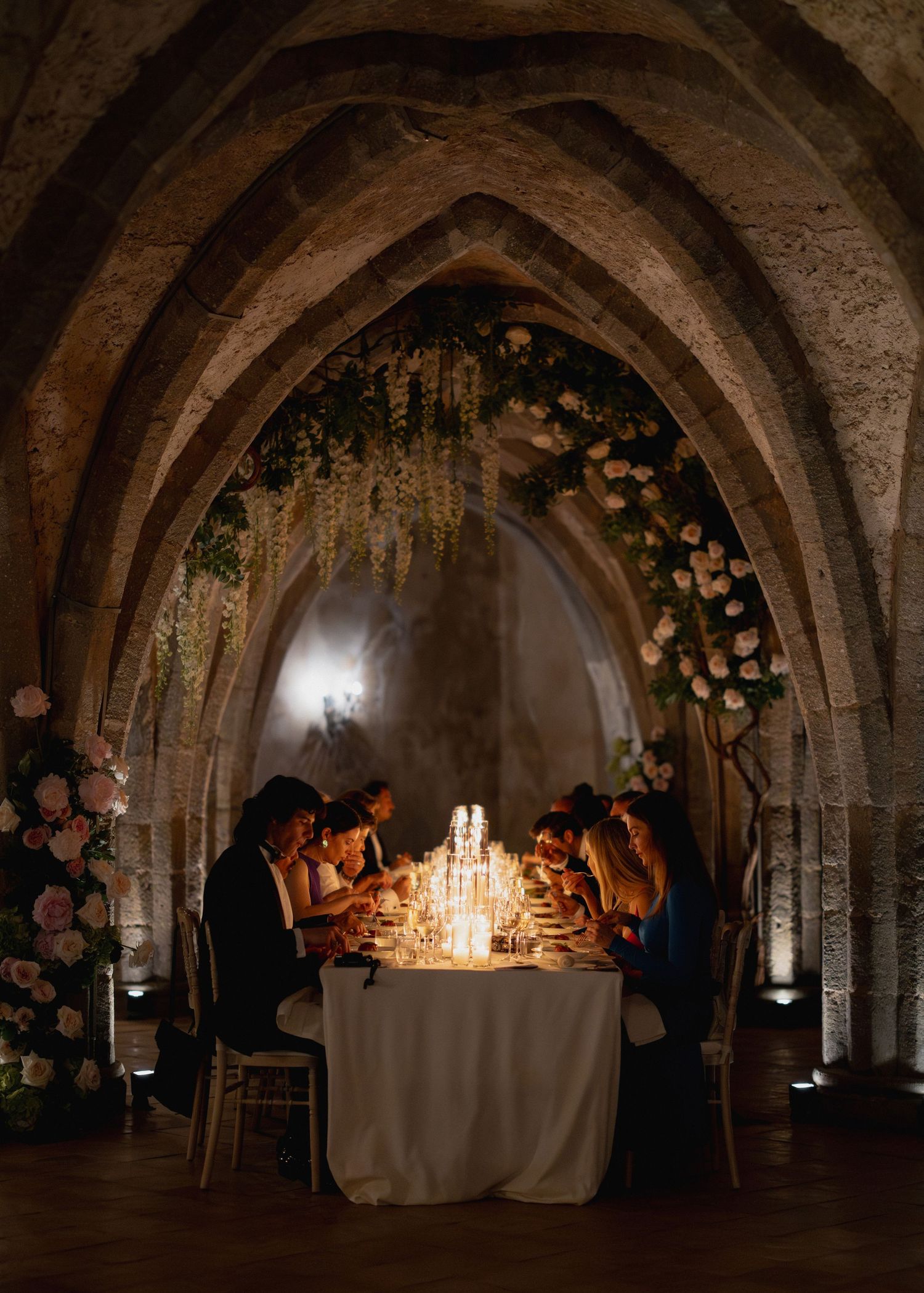 Guests dine by candlelight under gothic stone arches adorned with cascading flowers at an elegant wedding reception.