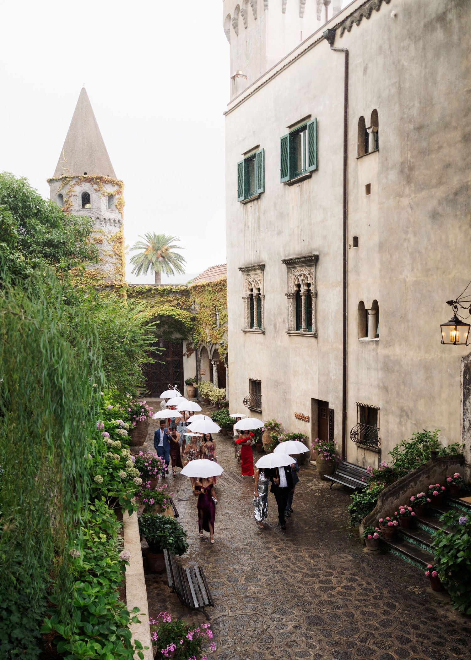 Guests walk with white umbrellas through a cobblestone courtyard beside a historic Italian villa on a rainy day.
