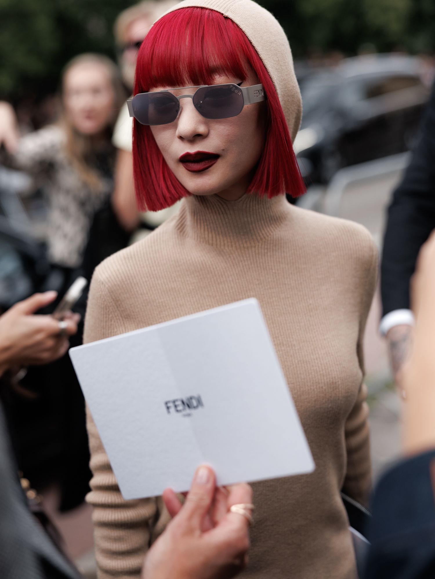 A person with bright red hair and sunglasses holds a white Fendi folder while wearing a beige turtleneck sweater.
