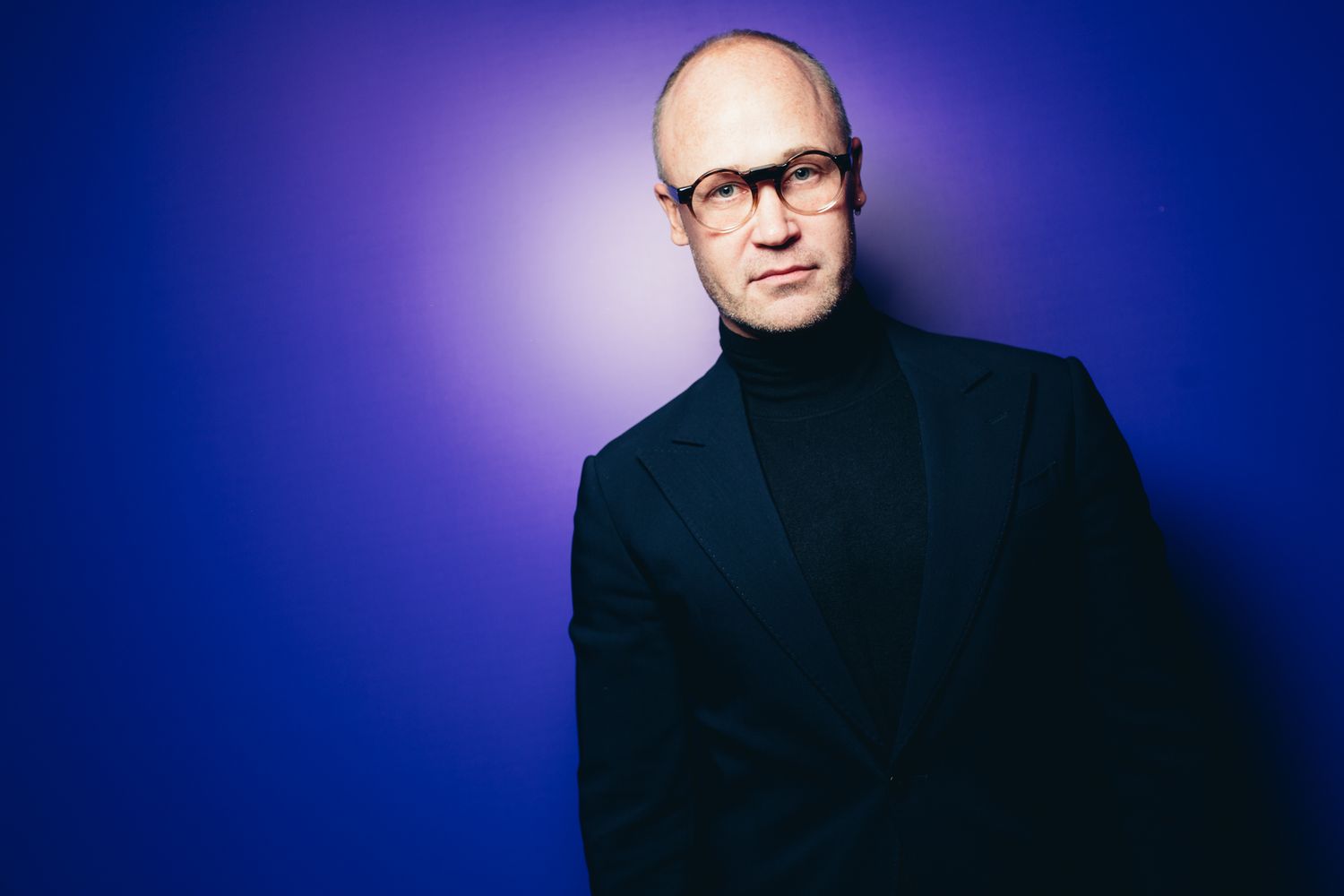 Portrait against blue backdrop showing person in black turtleneck and glasses.