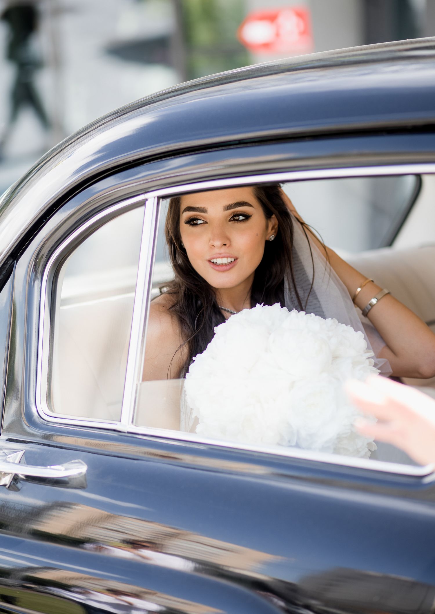 Bride in a classic black car holding a white bouquet, wearing a veil, ready for her wedding day.