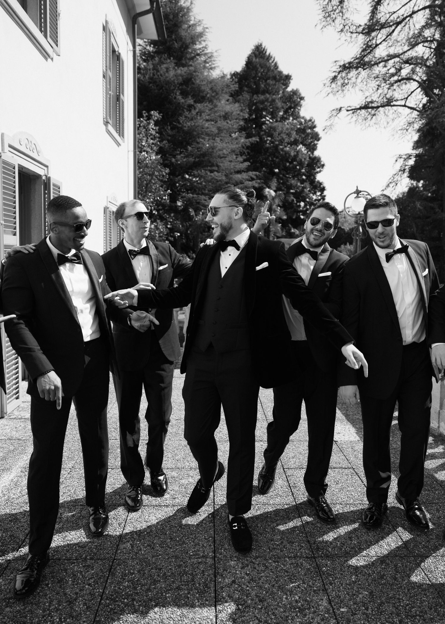 Black and white photo of five men in tuxedos with bow ties laughing and walking together outdoors.
