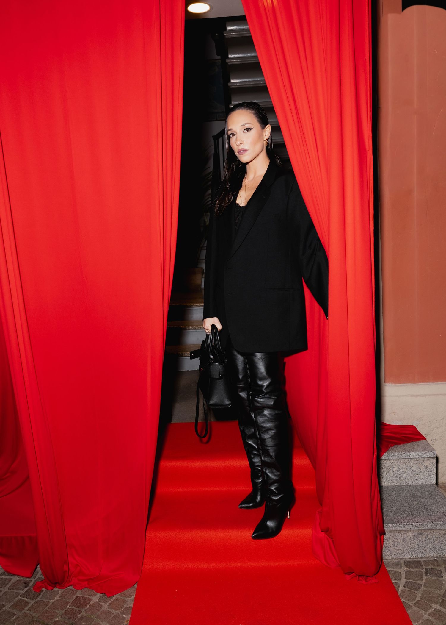 A stylish figure emerges through red curtains wearing black attire and knee-high boots on a red carpet.