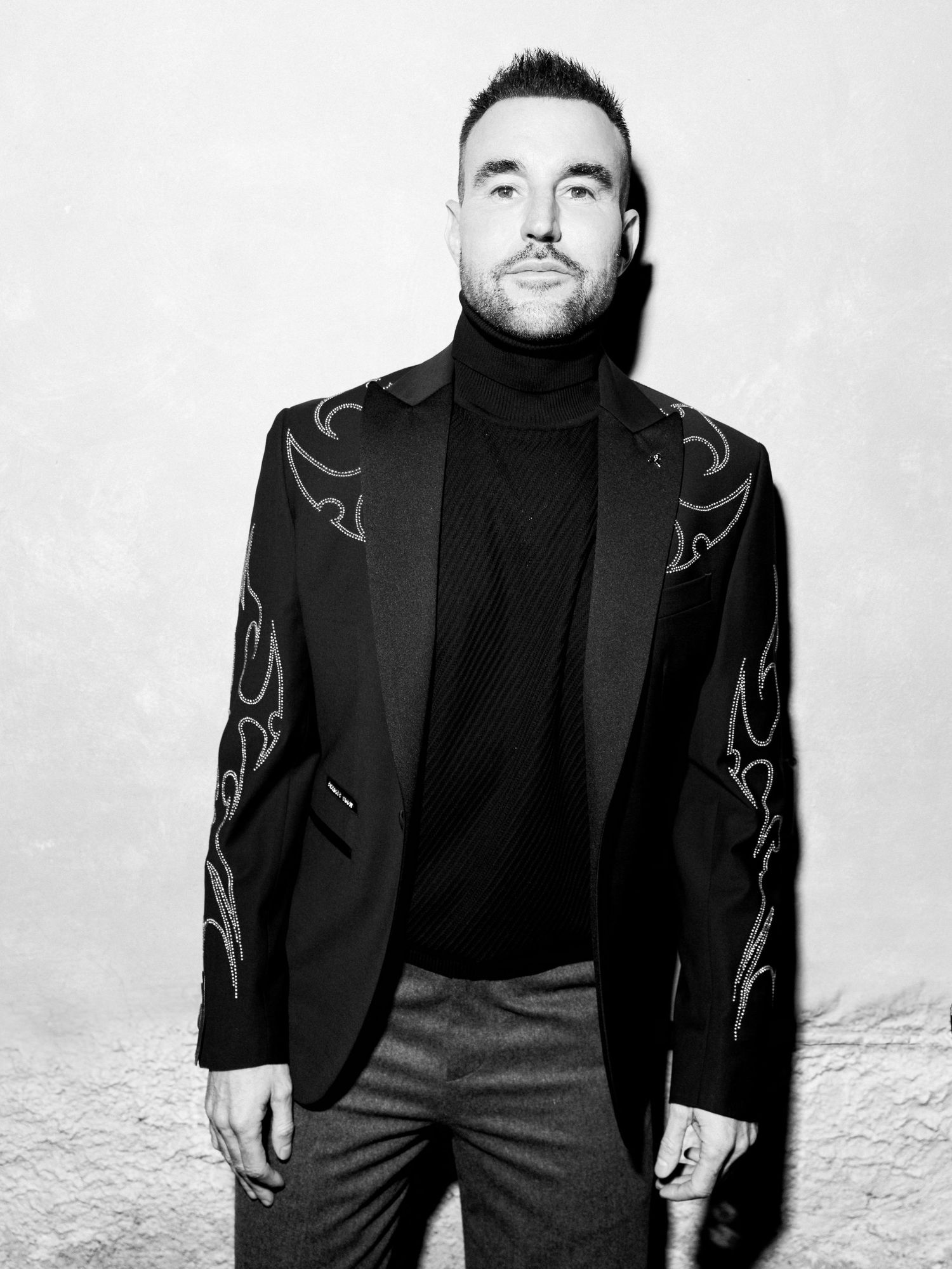 A person in a black turtleneck and blazer with embroidered details poses against a white background.