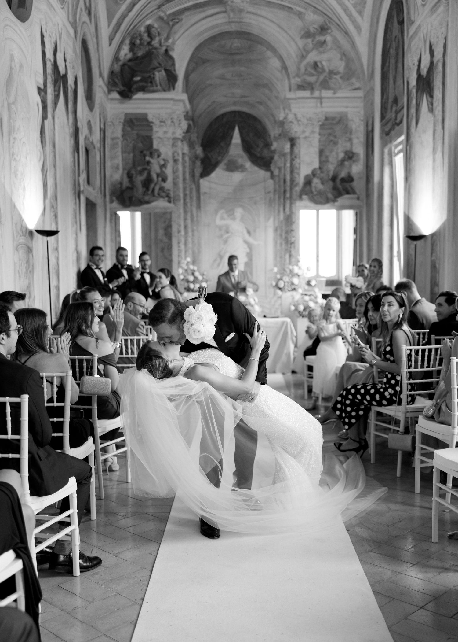 Black and white photo of bride and groom sharing a romantic dip kiss at elegant wedding ceremony in ornate venue.