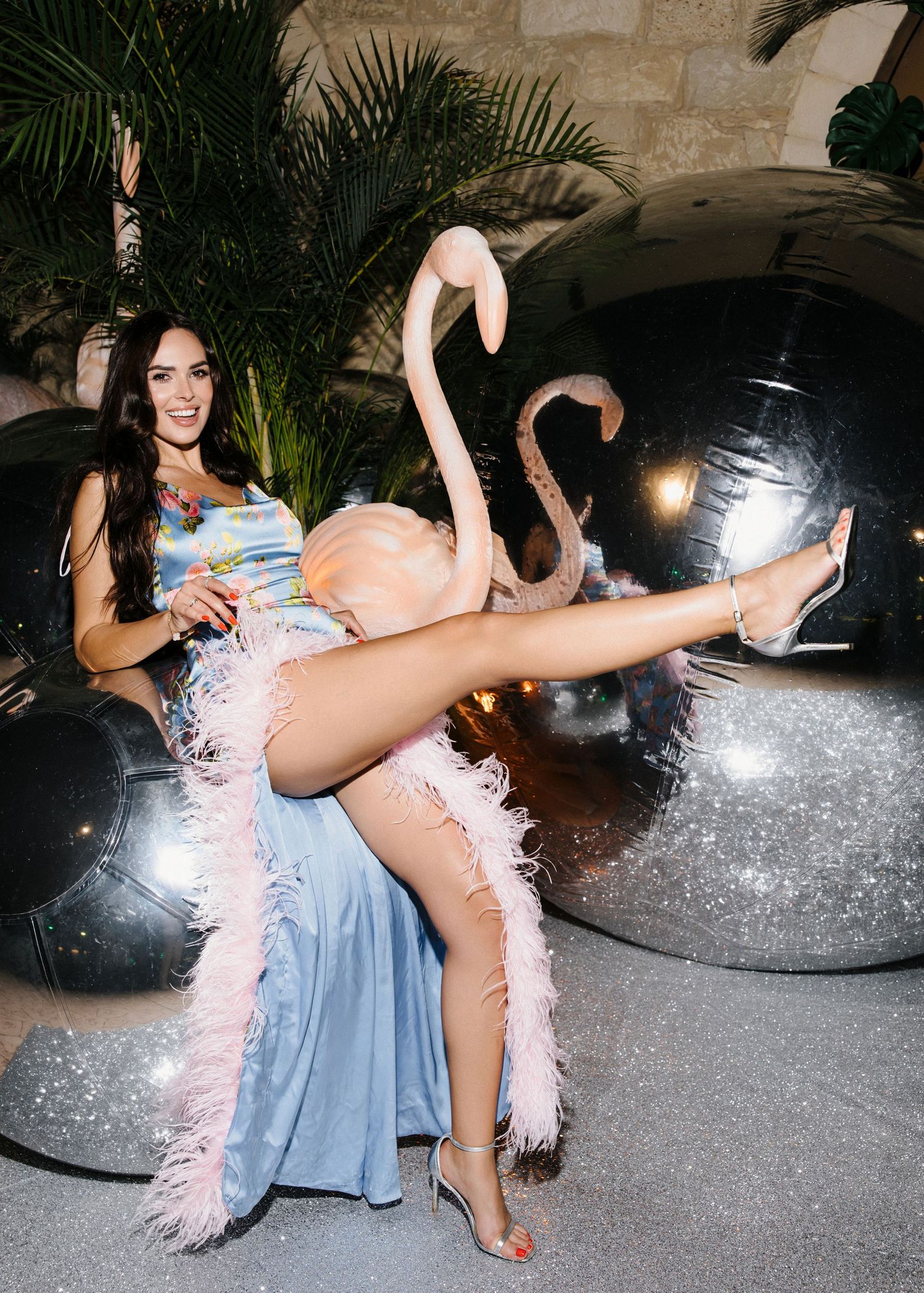 Woman in blue gown with pink feather boa poses playfully beside flamingo sculpture and metallic orbs at tropical party.