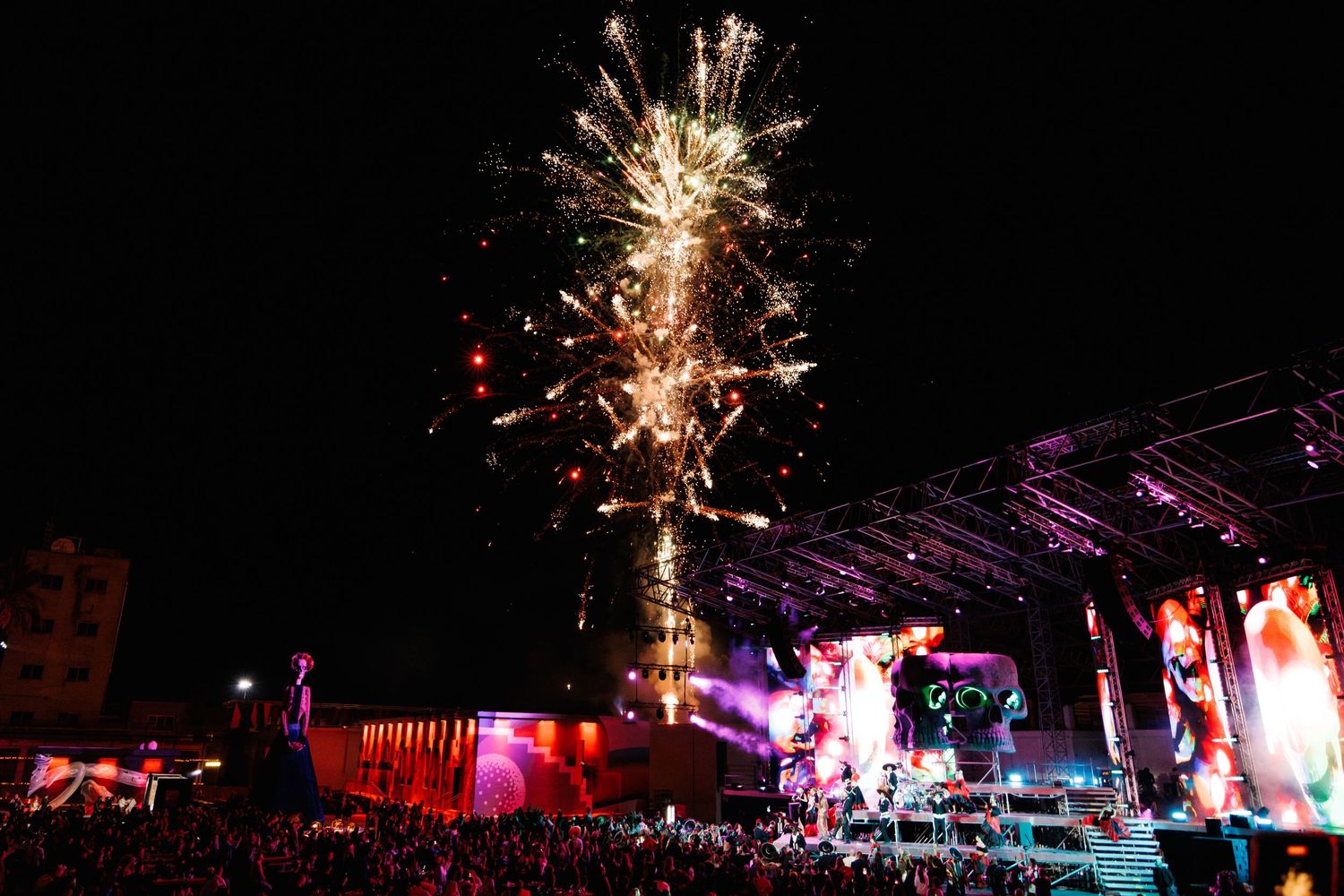 Fireworks exploding over an outdoor concert stage at night with colorful lighting.