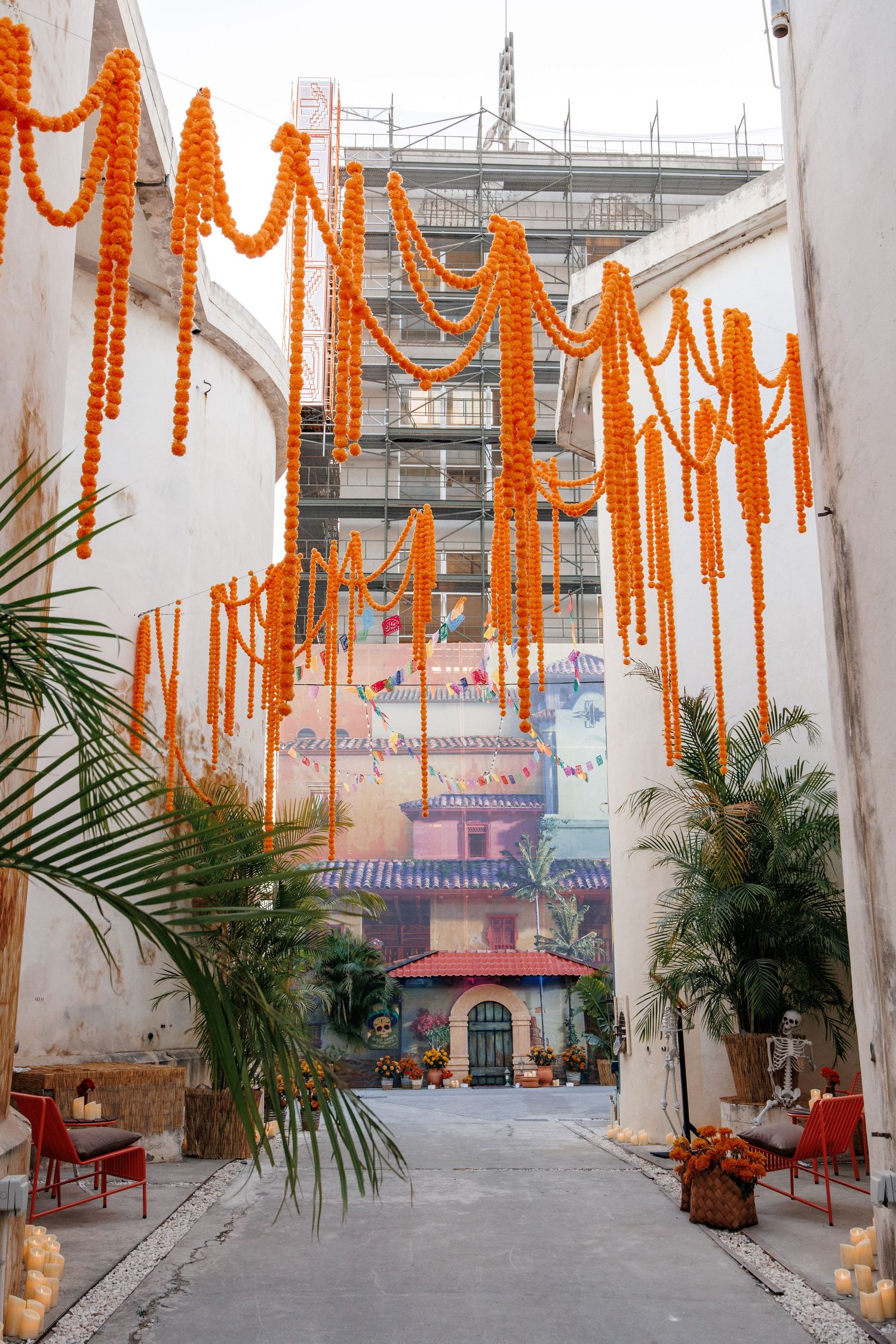 Orange marigold garlands hang between white walls in an alleyway decorated with palm trees and colorful artwork.