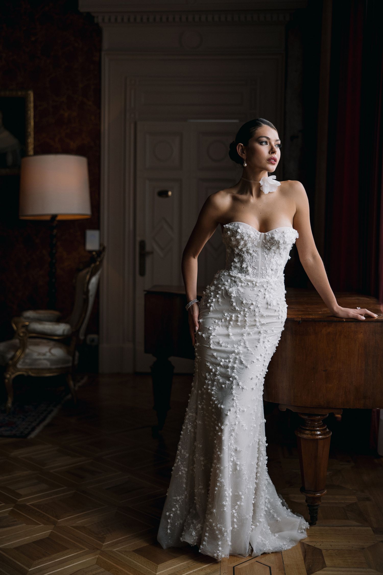 Dramatic bridal portrait in fitted lace gown against dark interior with vintage furniture.