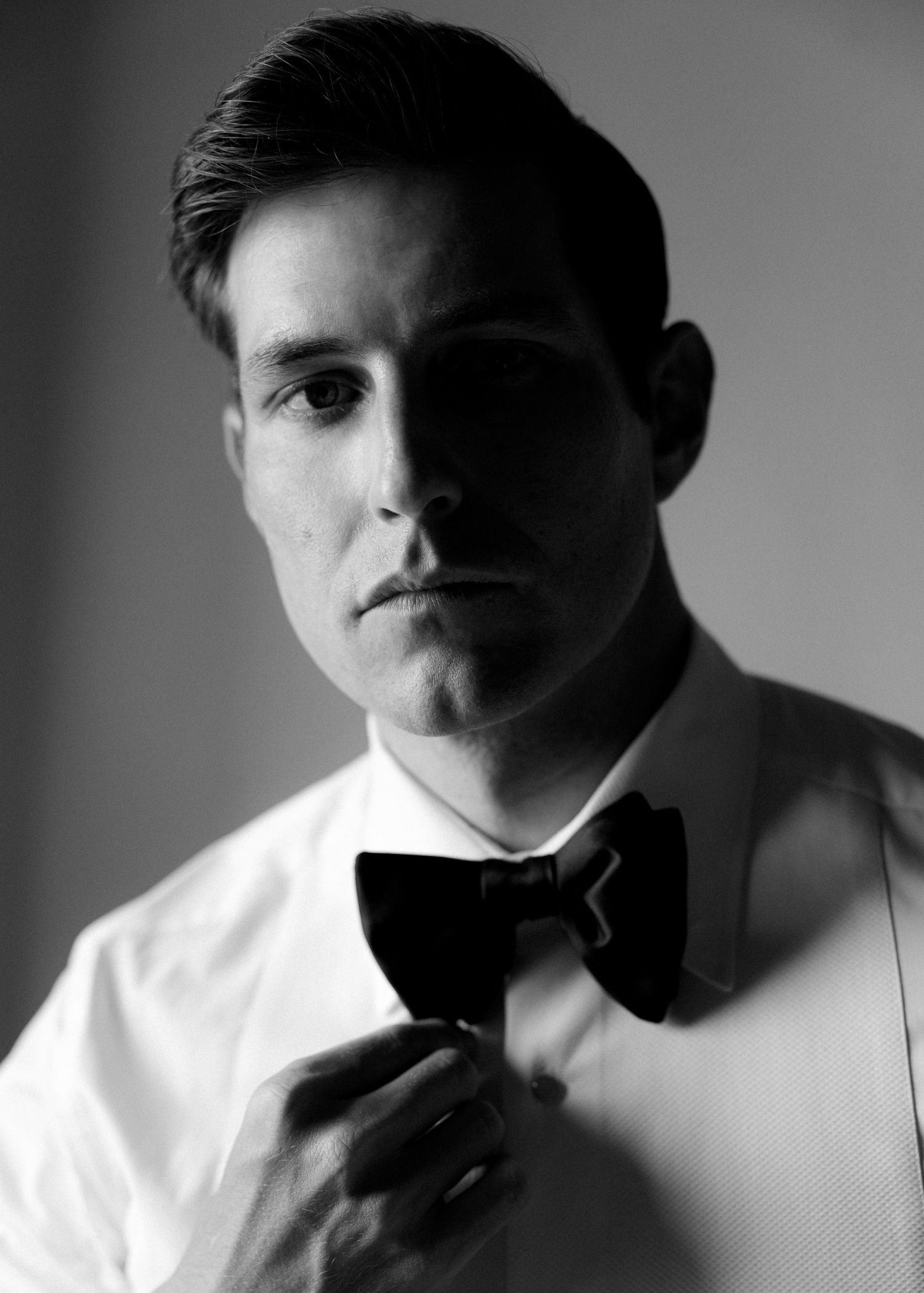 Black and white portrait of a young man in a white dress shirt adjusting his black bow tie.
