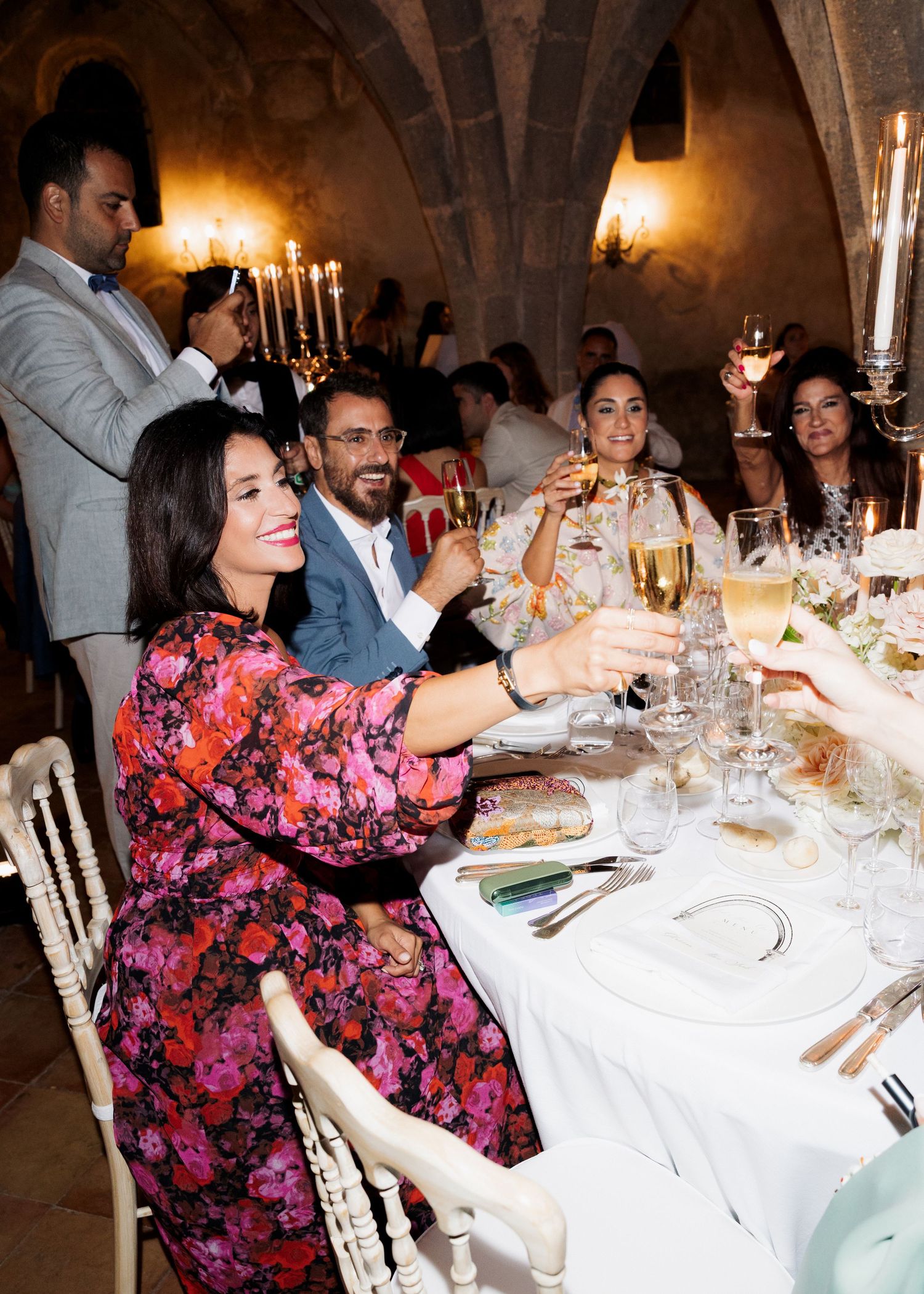 Guests in elegant attire raise champagne glasses at a candlelit dinner in a stone-walled venue.