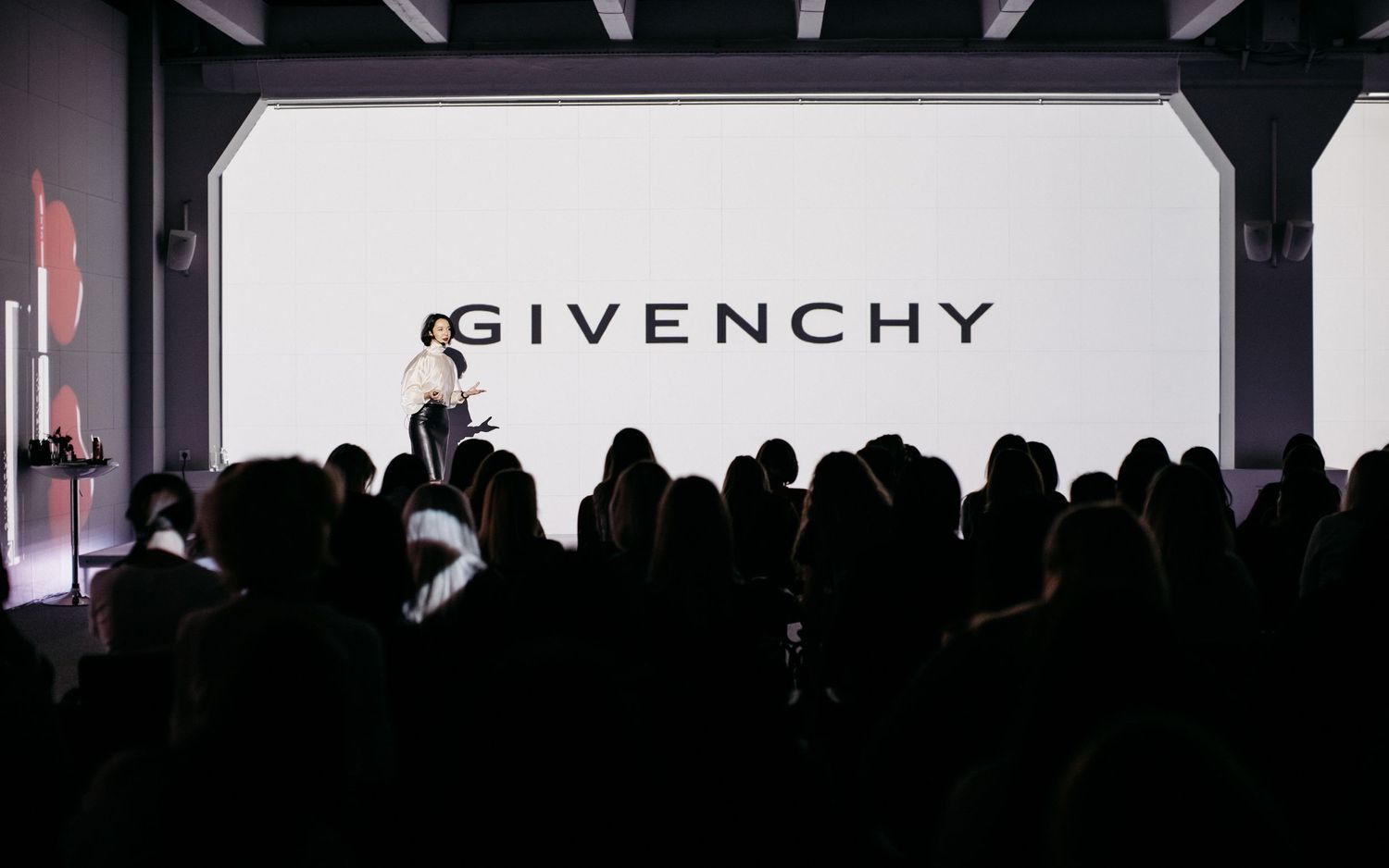 Silhouetted crowd watching Givenchy fashion show with illuminated white backdrop.